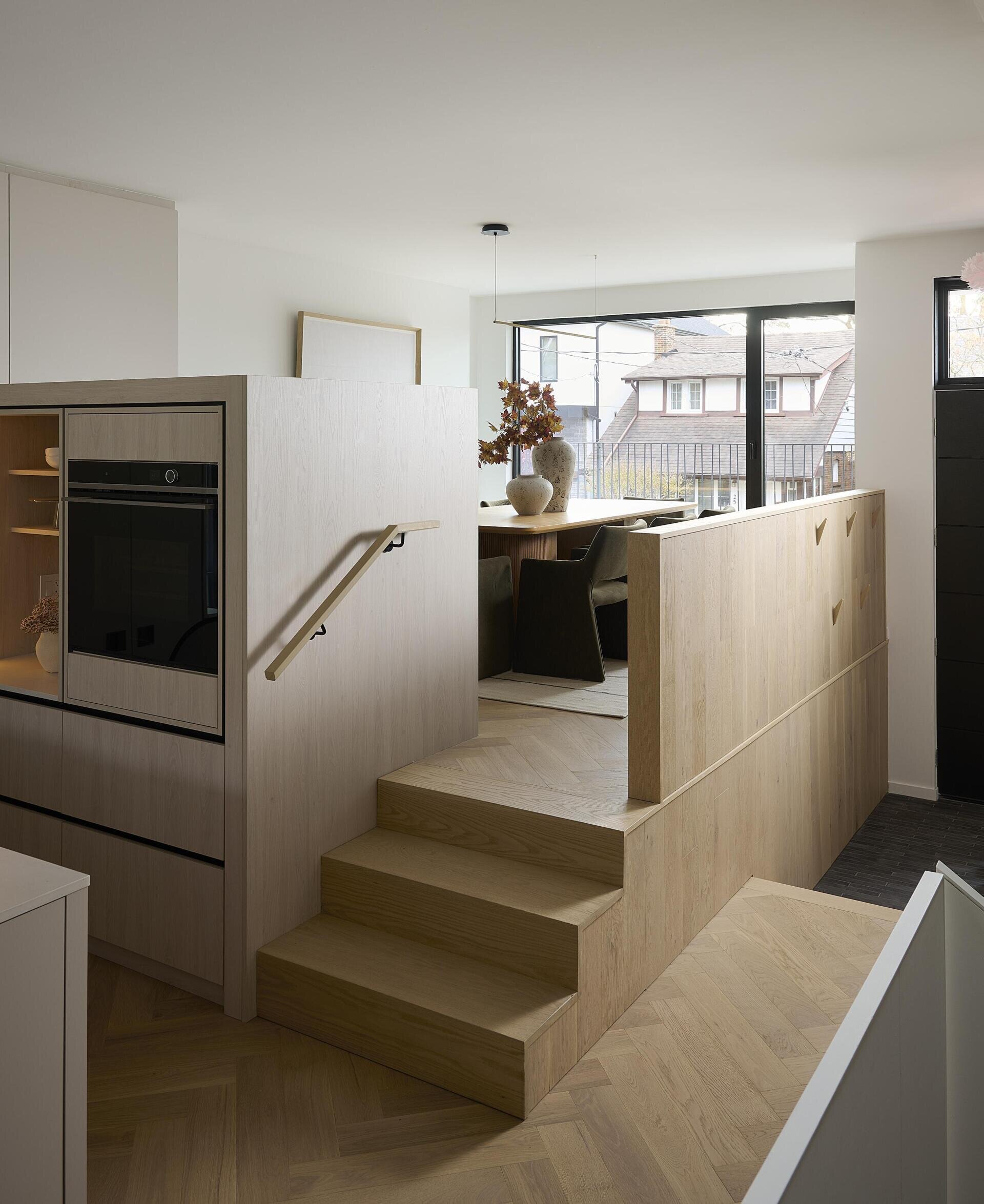 The dining area, accessed via a few steps, feels connected yet distinct, held by the warm tone of the oak cabinetry and the calm presence of the surrounding palette.