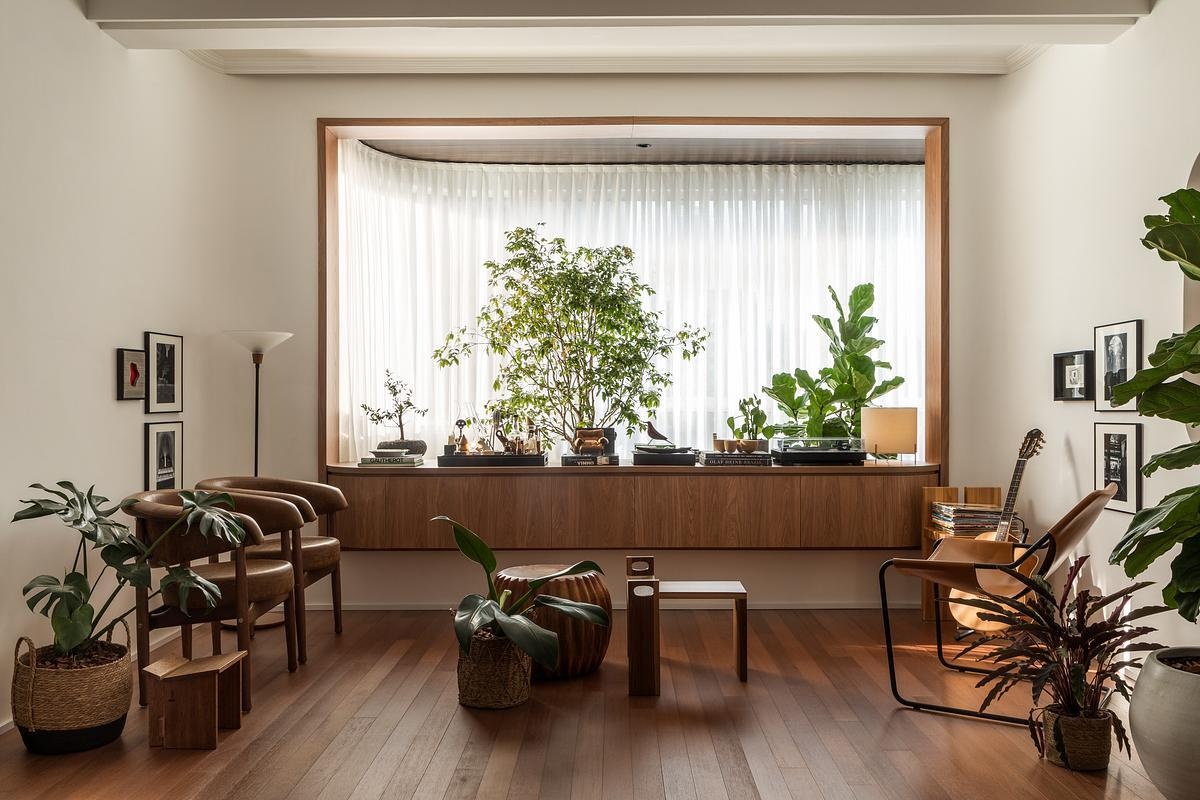 In a sitting area by a curved window, Brazilian design takes center stage. It centers on the Bag Armchair by Sérgio Rodrigues. Here, the chair sits near a Jabuticaba tree, creating a grounded, contemplative space. Personal photography, wood cabinetry, and a mix of Brazilian seating give it an intimate, collected atmosphere.