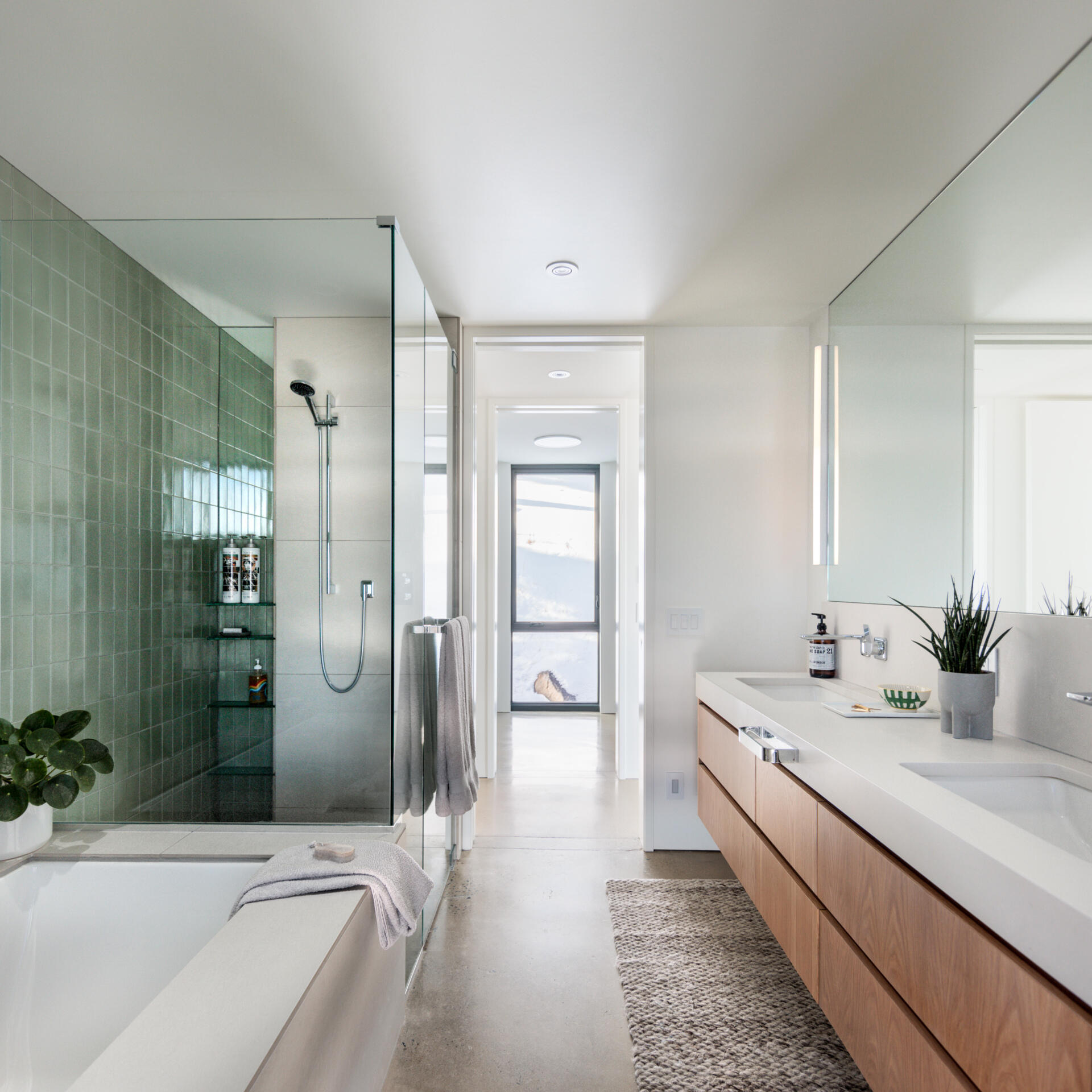 The bathroom stays true to the home’s quiet, efficient aesthetic. Soft green shower tiles, a simple glass enclosure, and a long floating vanity keep the space calm and bright, while southern light warms the room naturally.