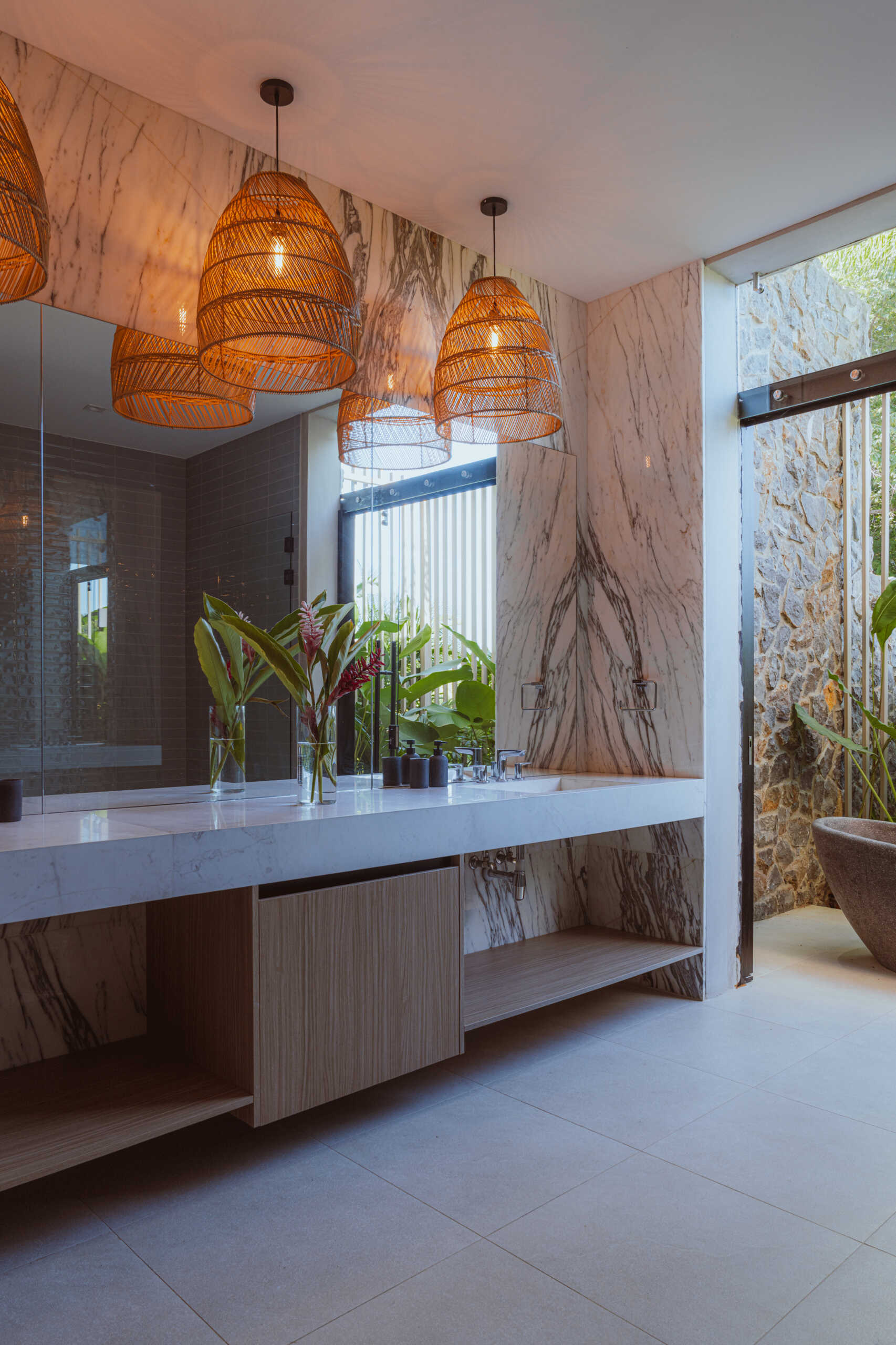 The bathroom features a long vanity topped with woven pendant lights that add warmth and texture. A freestanding bathtub sits nearby, surrounded by lush plants, creating a serene, spa-like corner that connects the space to the home’s natural surroundings.