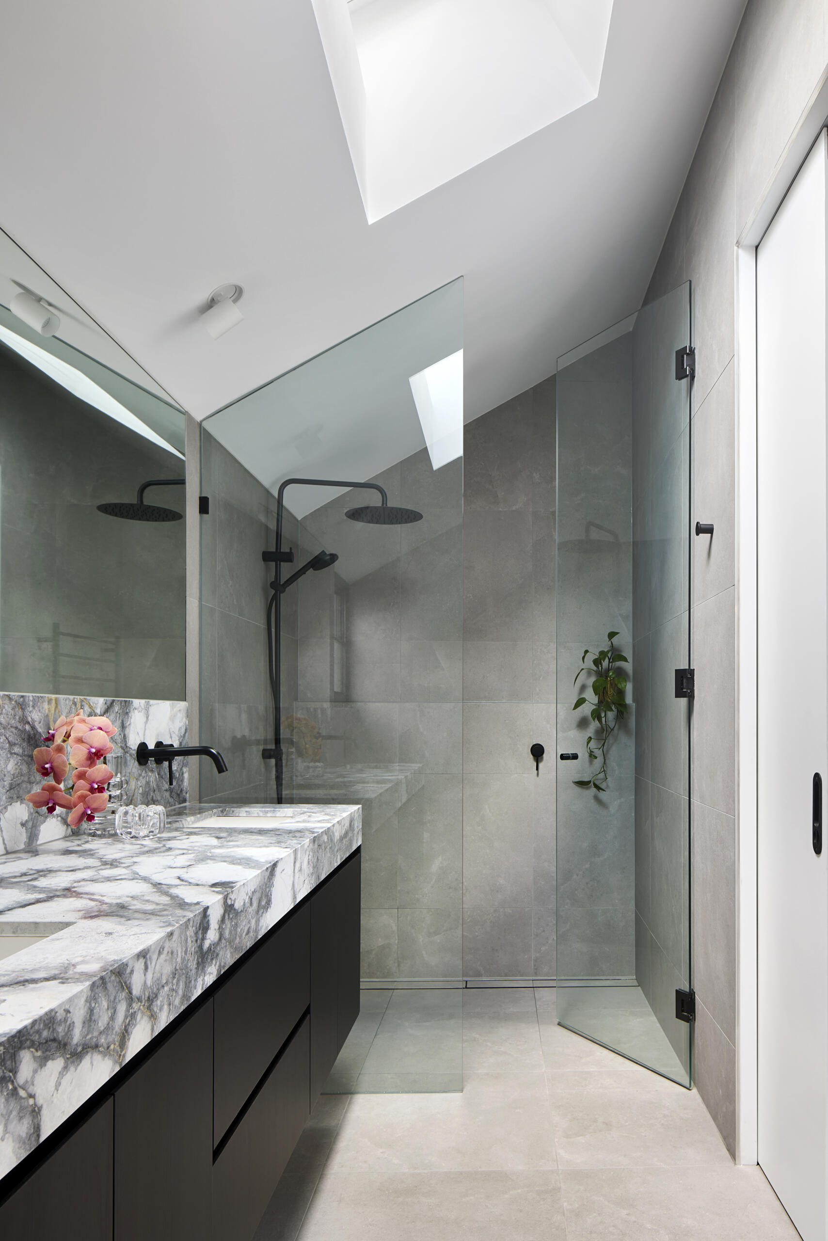 The bathroom feels calm and restrained, shaped by soft grey tiles and a clear frameless shower. Light from the skylight lifts the space and highlights the veining in the marble countertop. A trailing plant and a small touch of pink soften the palette, giving the room a quiet sense of ease.