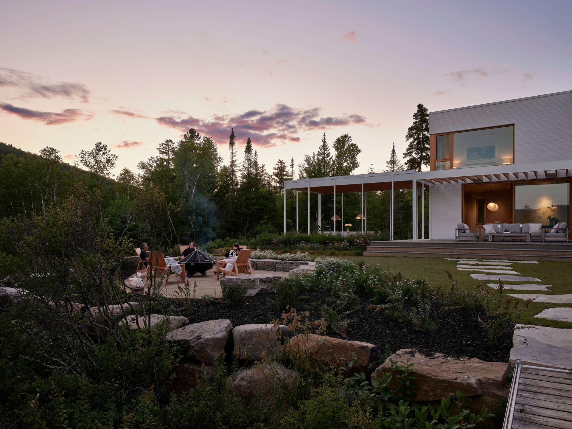 A stone path leads from the veranda through native plantings toward a fire pit and a sandy beach. 