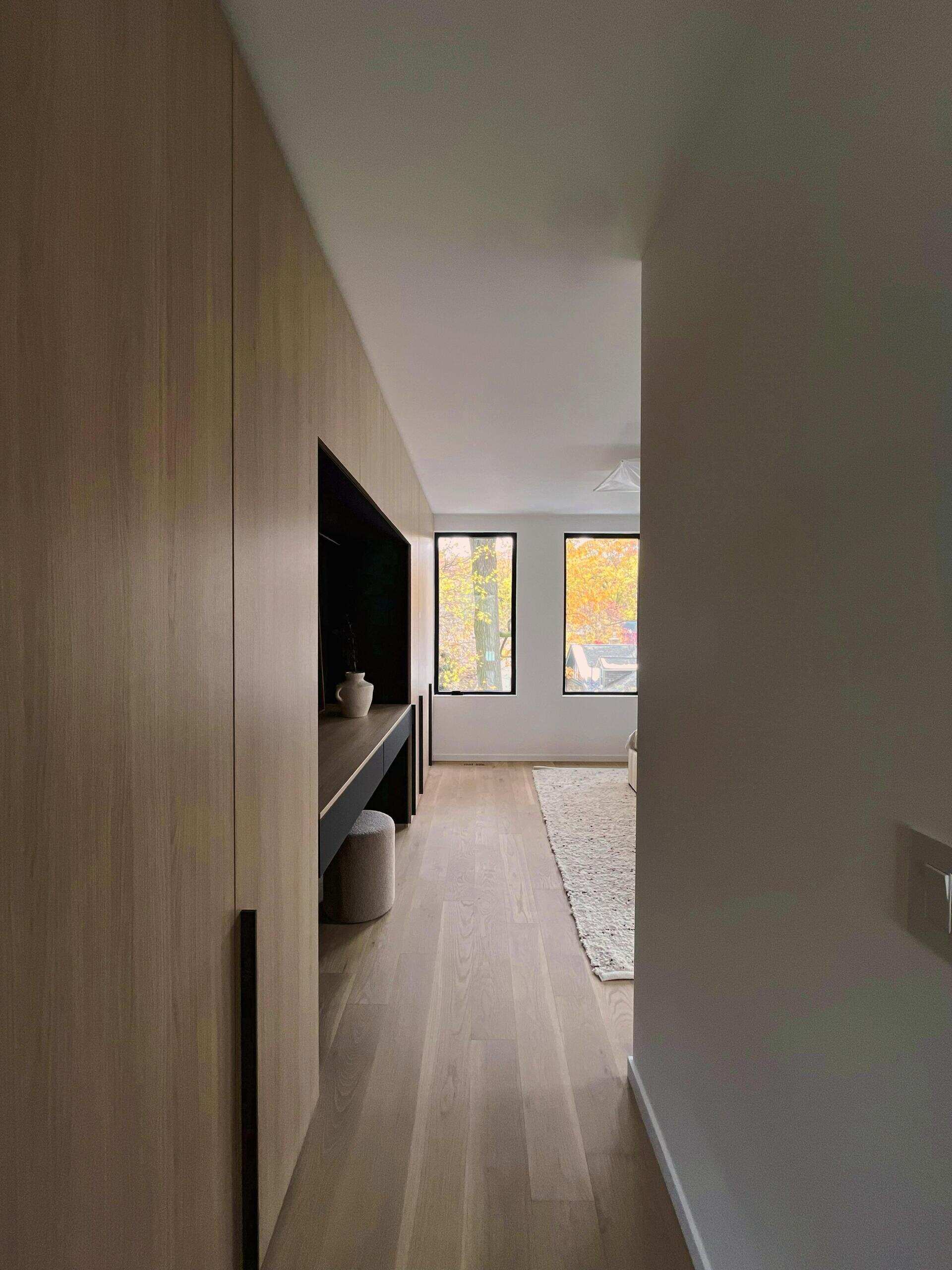 Located off the family room, the principal bedroom uses windows to frame treetop views. Along one wall, quarter cut oak and matte graphite built-ins integrate wardrobe space, a vanity, and a concealed door to a flexible room that can become a hidden office or extended storage.