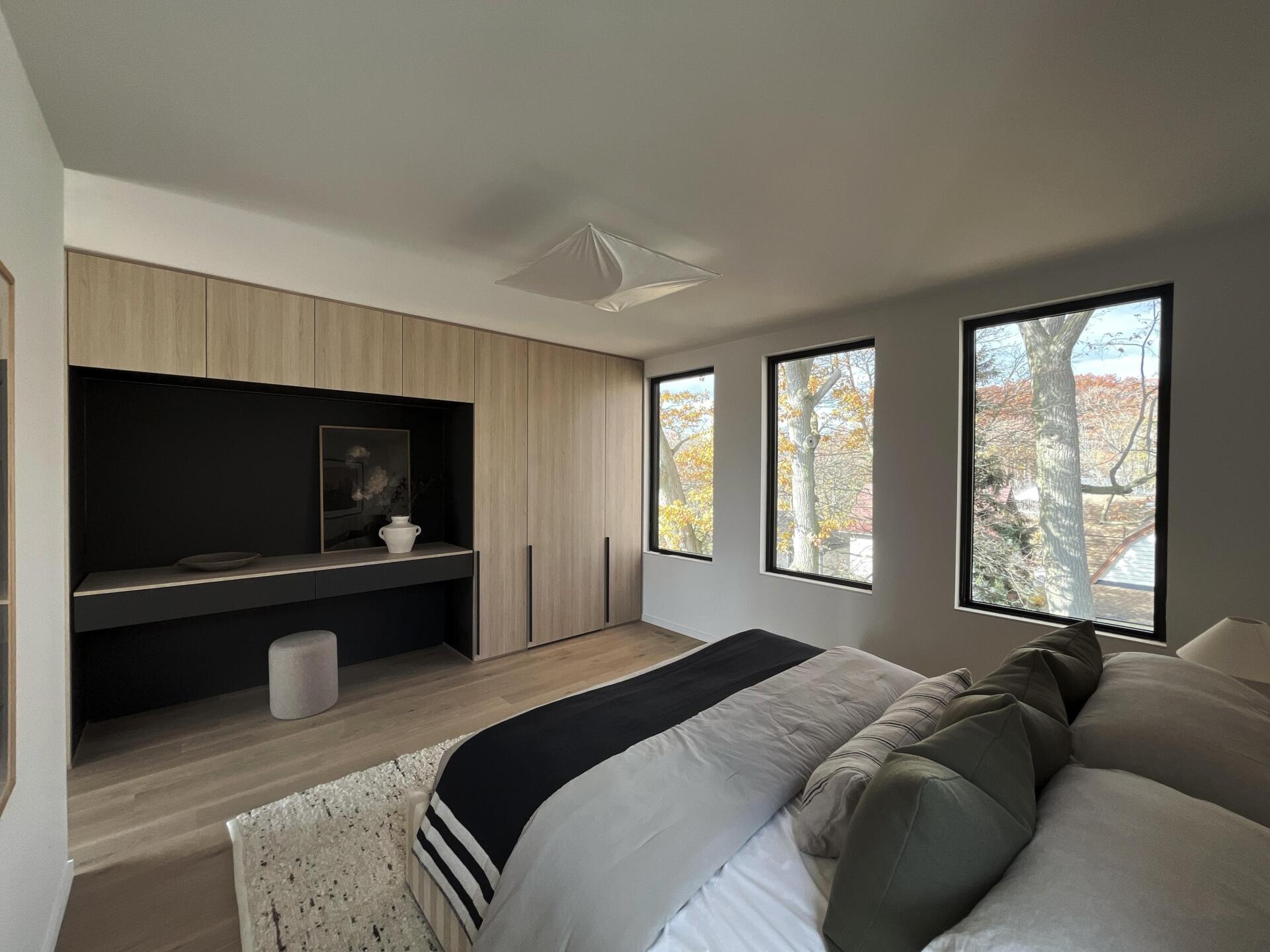 Located off the family room, the principal bedroom uses windows to frame treetop views. Along one wall, quarter cut oak and matte graphite built-ins integrate wardrobe space, a vanity, and a concealed door to a flexible room that can become a hidden office or extended storage.