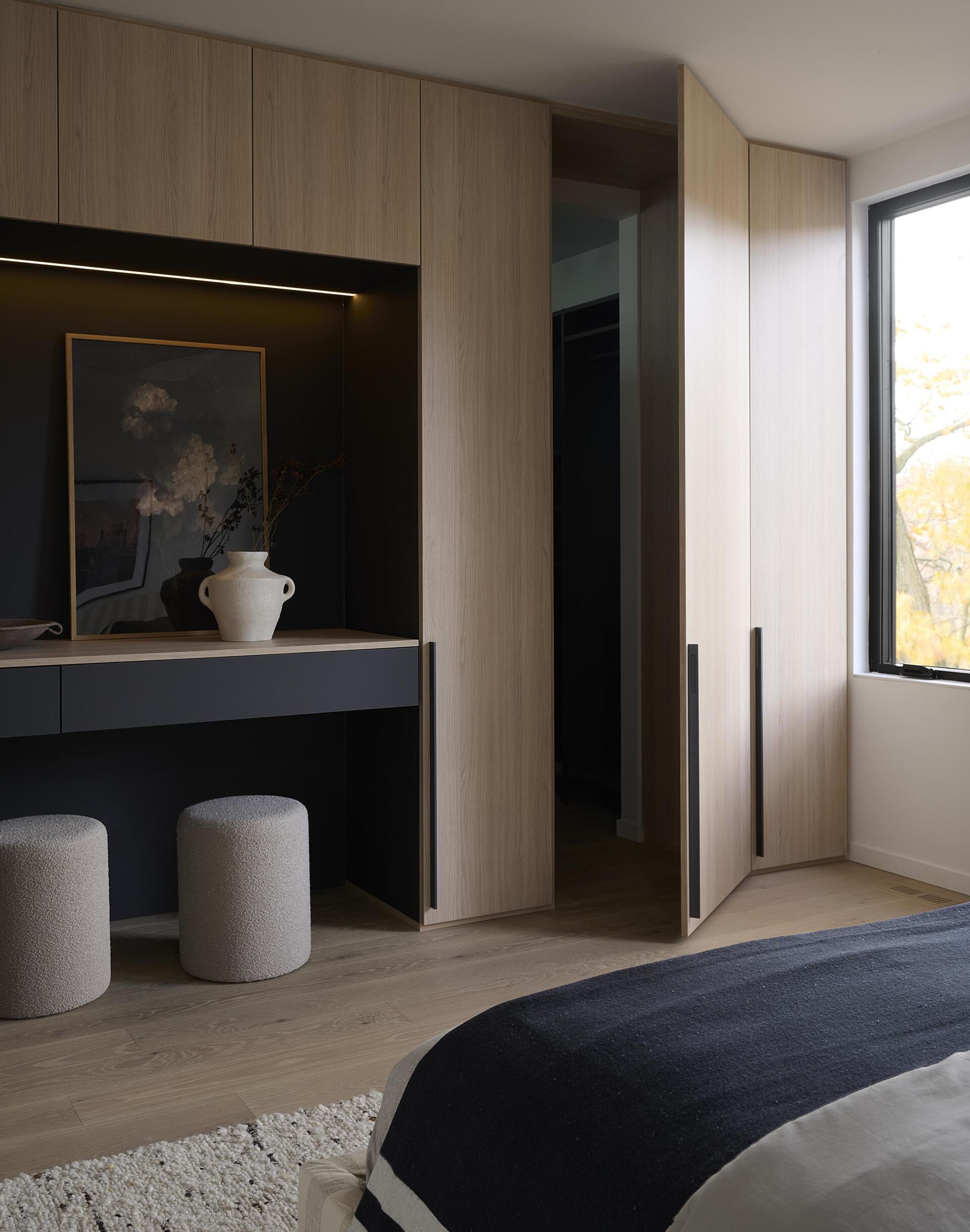 Located off the family room, the principal bedroom uses windows to frame treetop views. Along one wall, quarter cut oak and matte graphite built-ins integrate wardrobe space, a vanity, and a concealed door to a flexible room that can become a hidden office or extended storage.