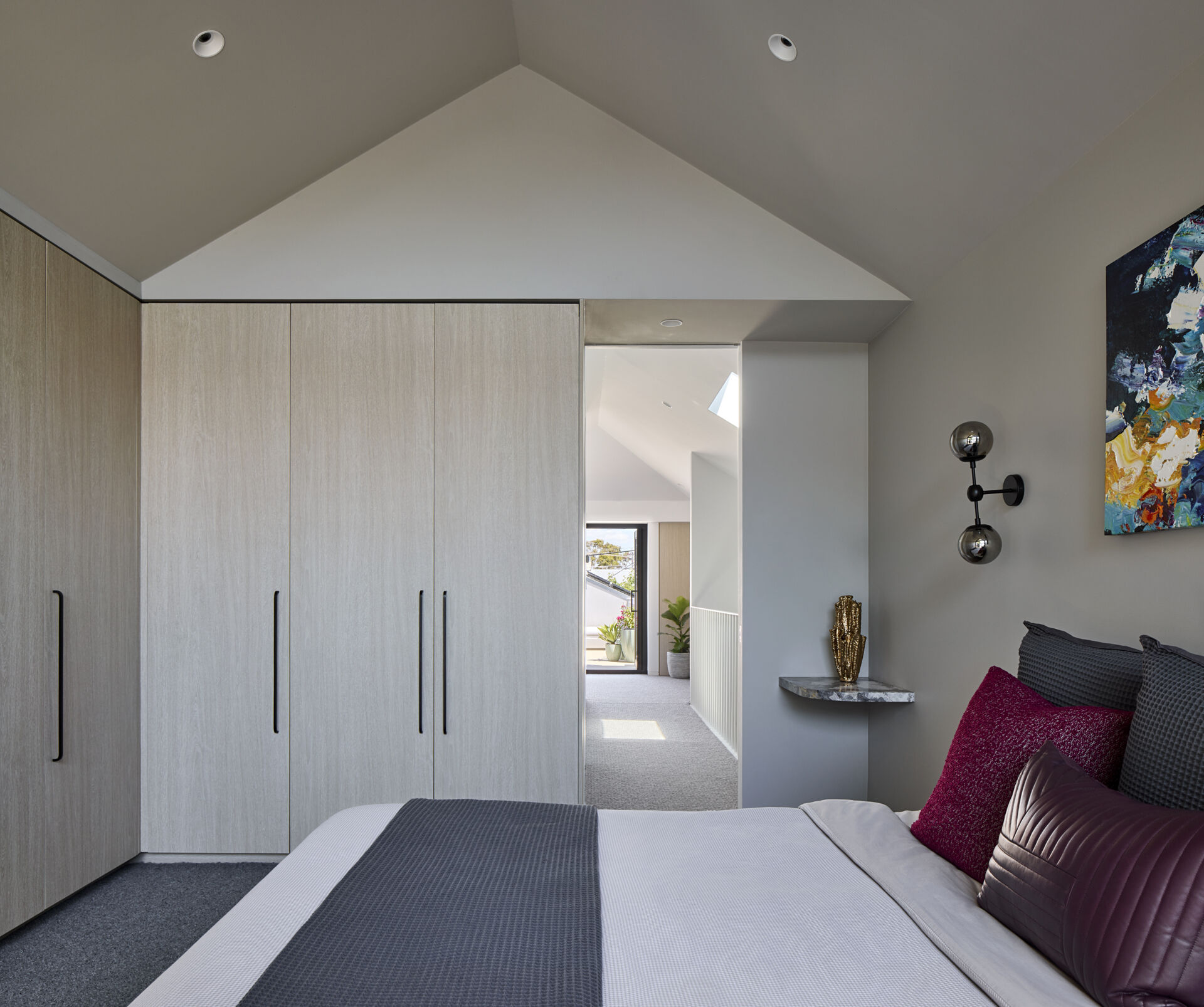 In this bedroom, there's a gentle, tailored feel with built-in pale wood closets. Light filters through from the hallway and settles across the textured carpet and crisp bedding. Plum pillows and a simple artwork add a measured hint of color while the small marble shelf and sculptural light keep the room tidy and restful.