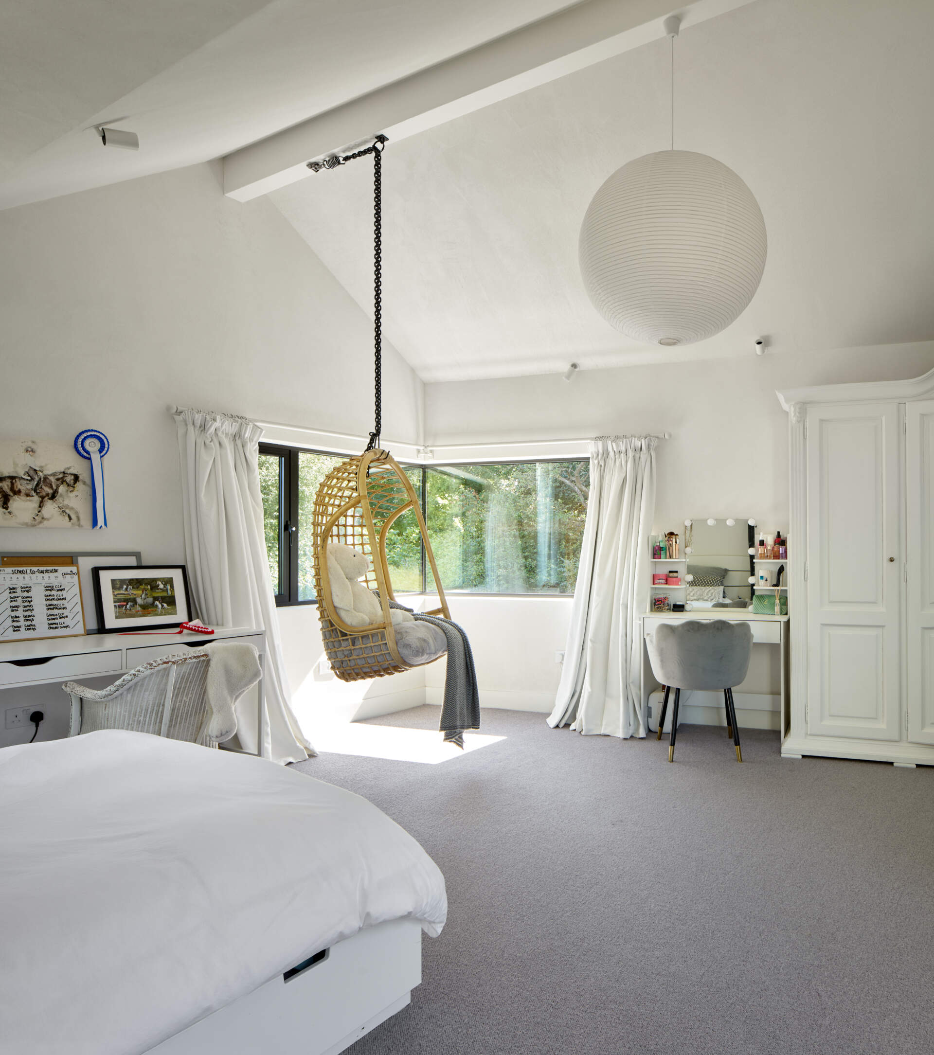 This bedroom is shaped by soft light and simple comfort. Each space feels open and calm, with tall ceilings that lift the room and windows that frame the surrounding greenery. Details stay personal and practical, from the built in storage to the small vanity tucked beside the curtains.