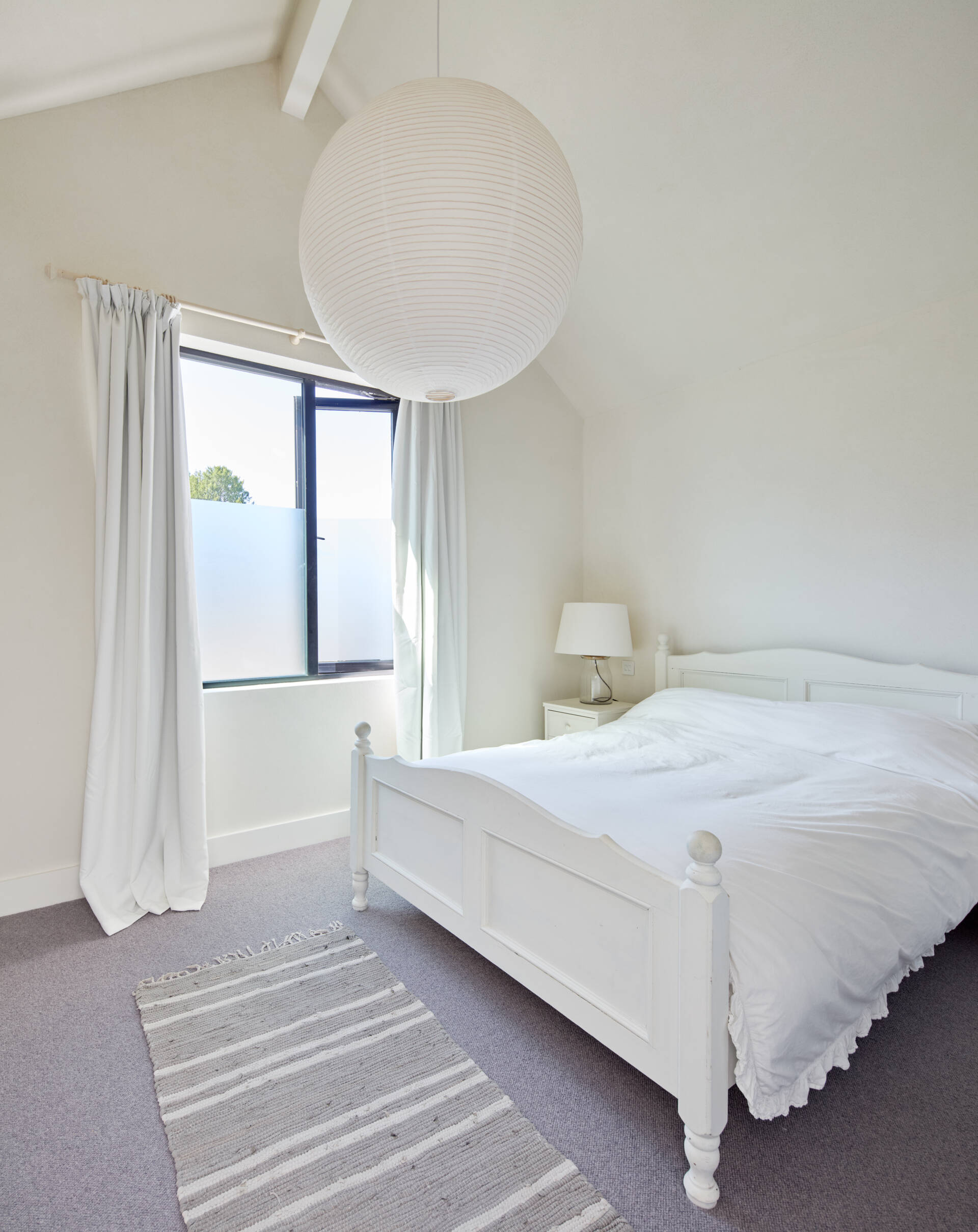 This bedroom is shaped by soft light and simple comfort. Each space feels open and calm, with tall ceilings that lift the room and windows that frame the surrounding greenery. Details stay personal and practical, from the built in storage to the small vanity tucked beside the curtains.