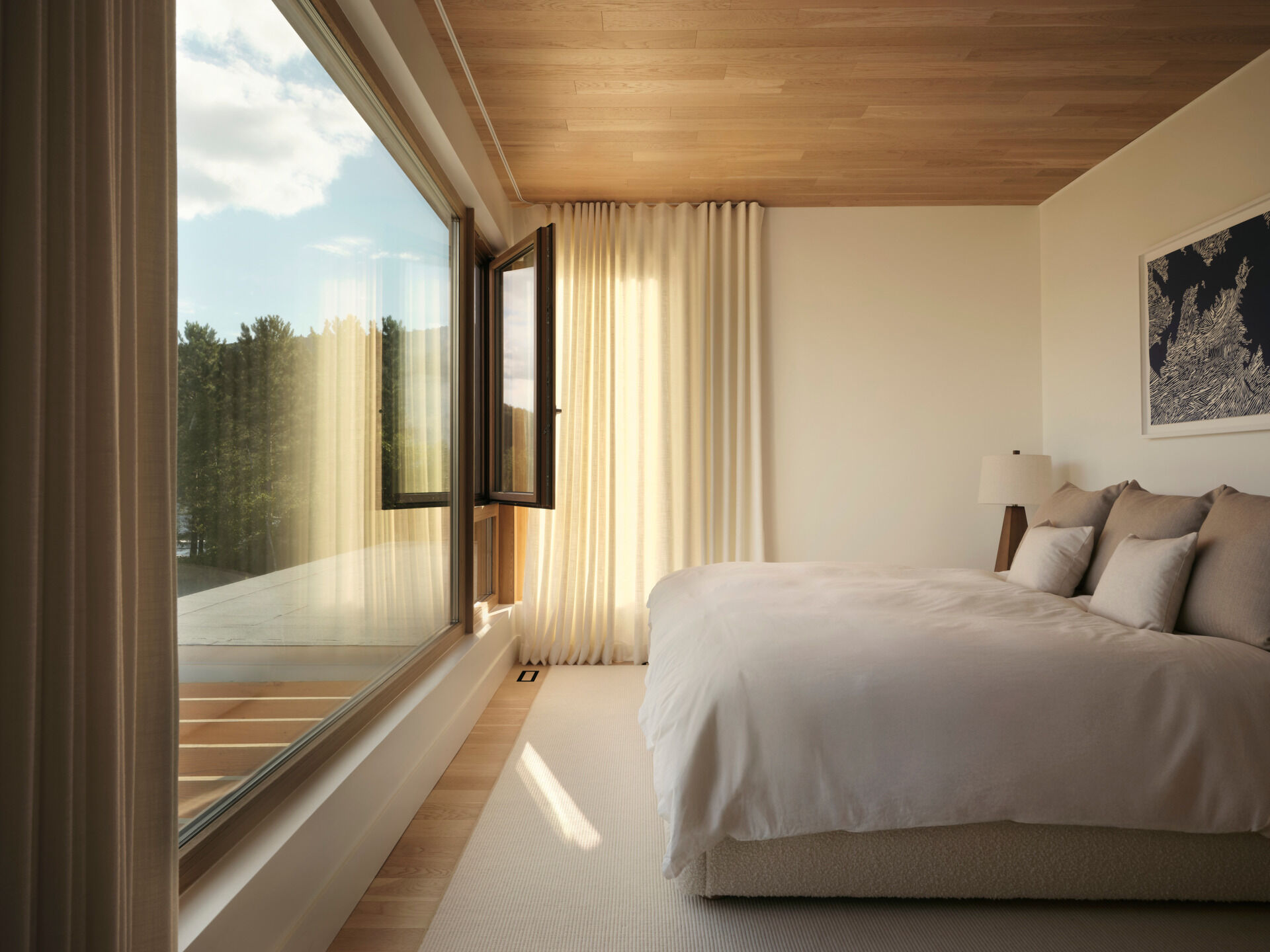 In this bedroom, soft linens, pale walls, and oak accents frame the view of the outdoors beyond the picture window. There are no distractions, only calm textures and quiet light.