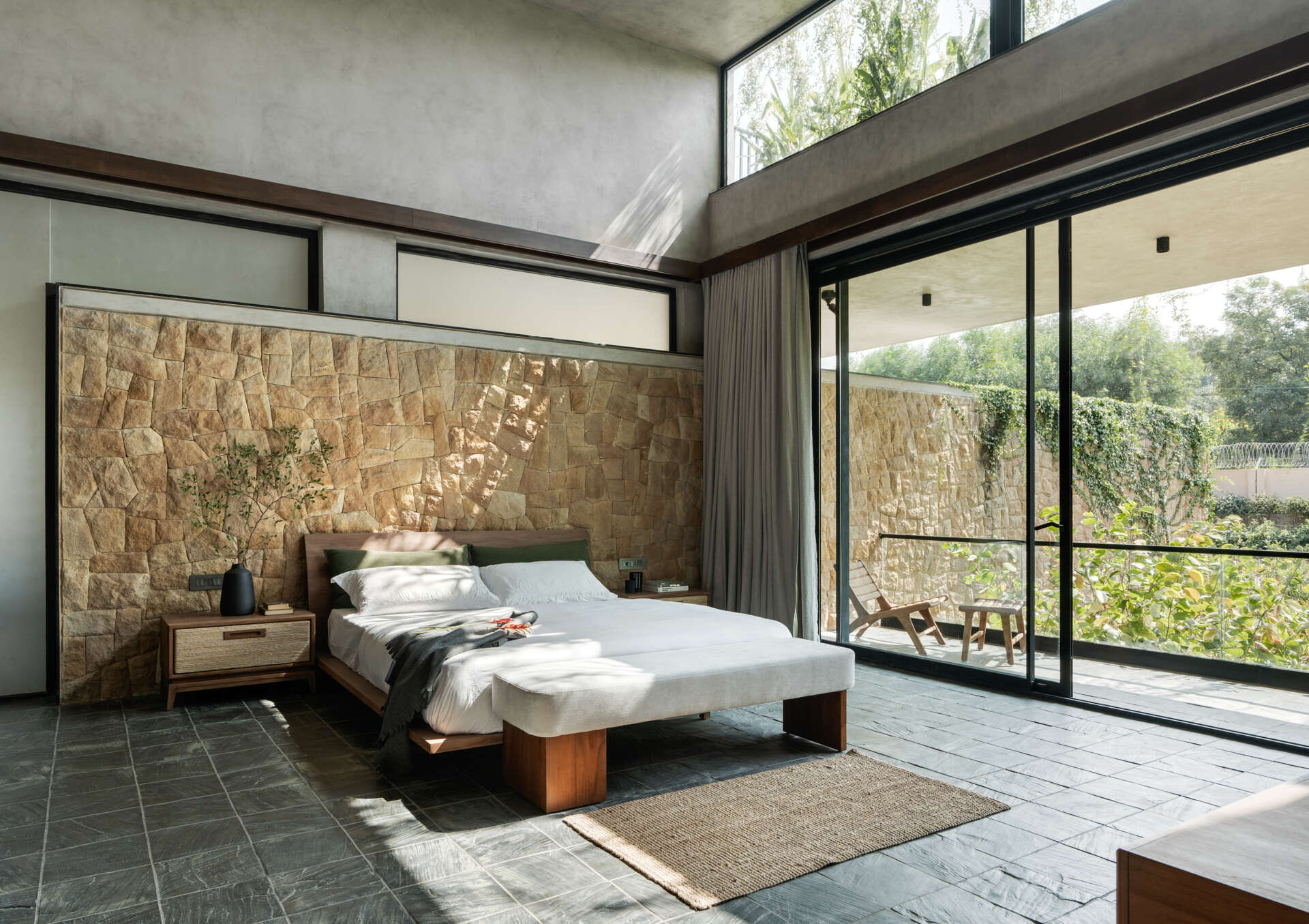 A modern bedroom framed by stone walls and large windows that welcome in light and greenery.