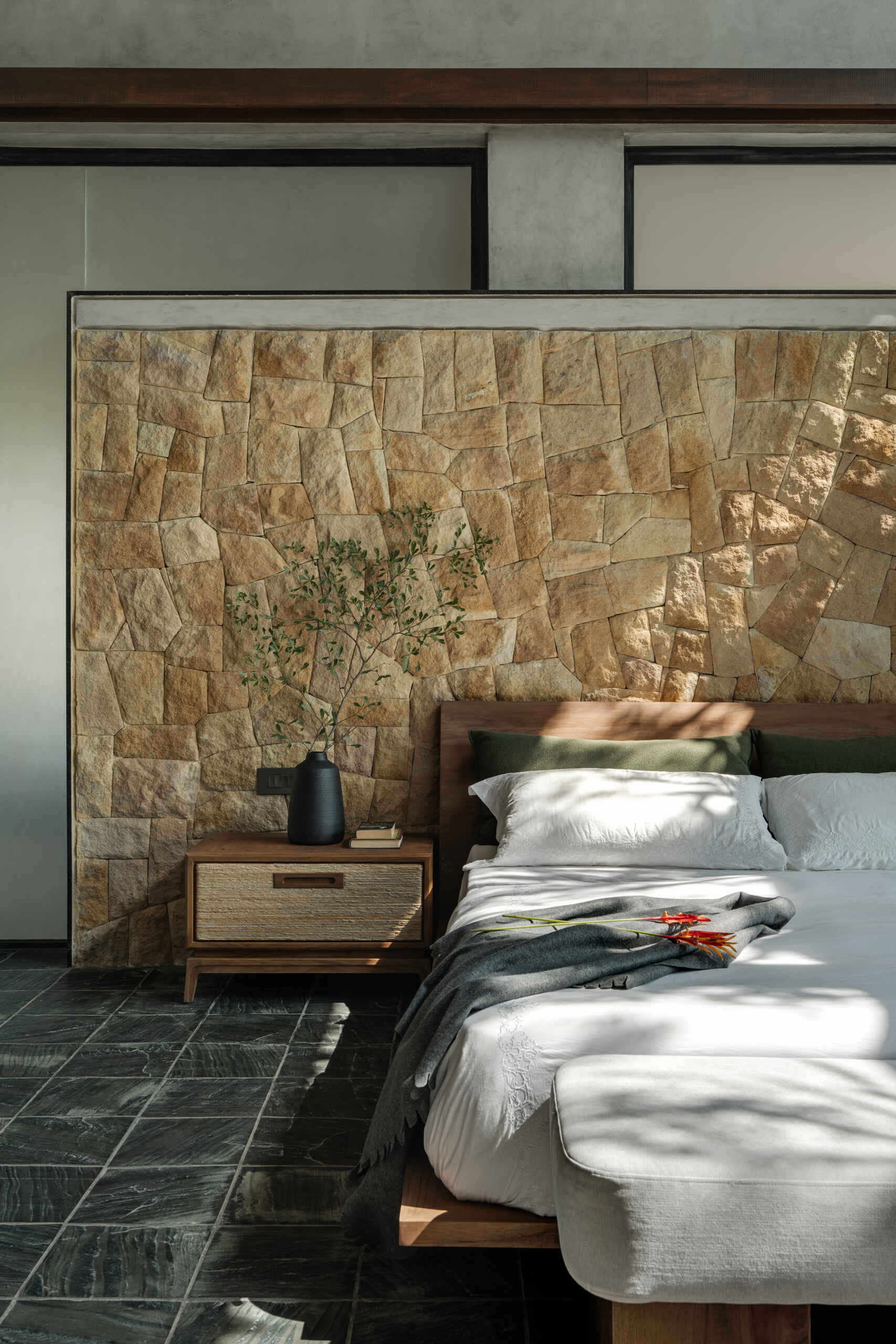 A modern bedroom framed by stone walls and large windows that welcome in light and greenery.