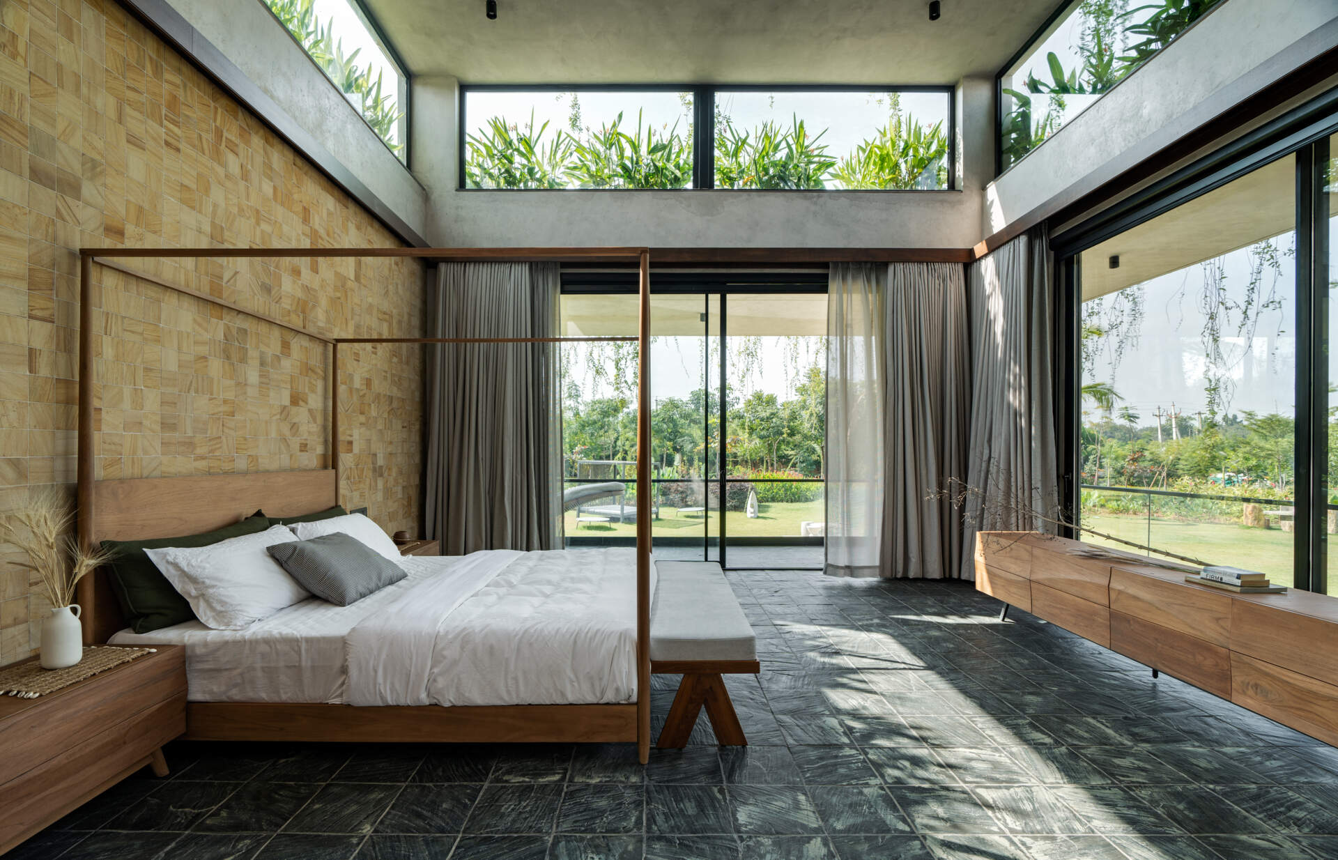 A modern bedroom framed by stone walls and large windows that welcome in light and greenery.