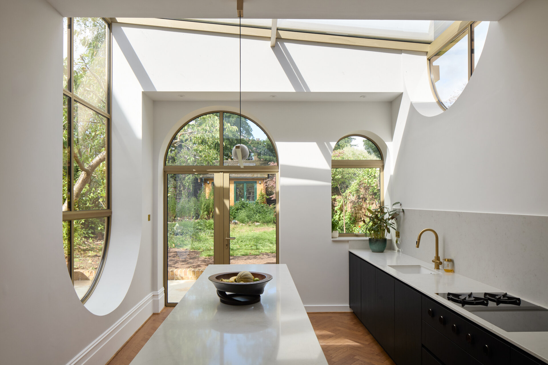 In the kitchen of Perry Vale House, inverted arches and a wraparound glass roof fill the space with natural light. Soft grey cabinetry, marble worktops, and timber tones create a warm, modern feel that stays true to the home’s character.