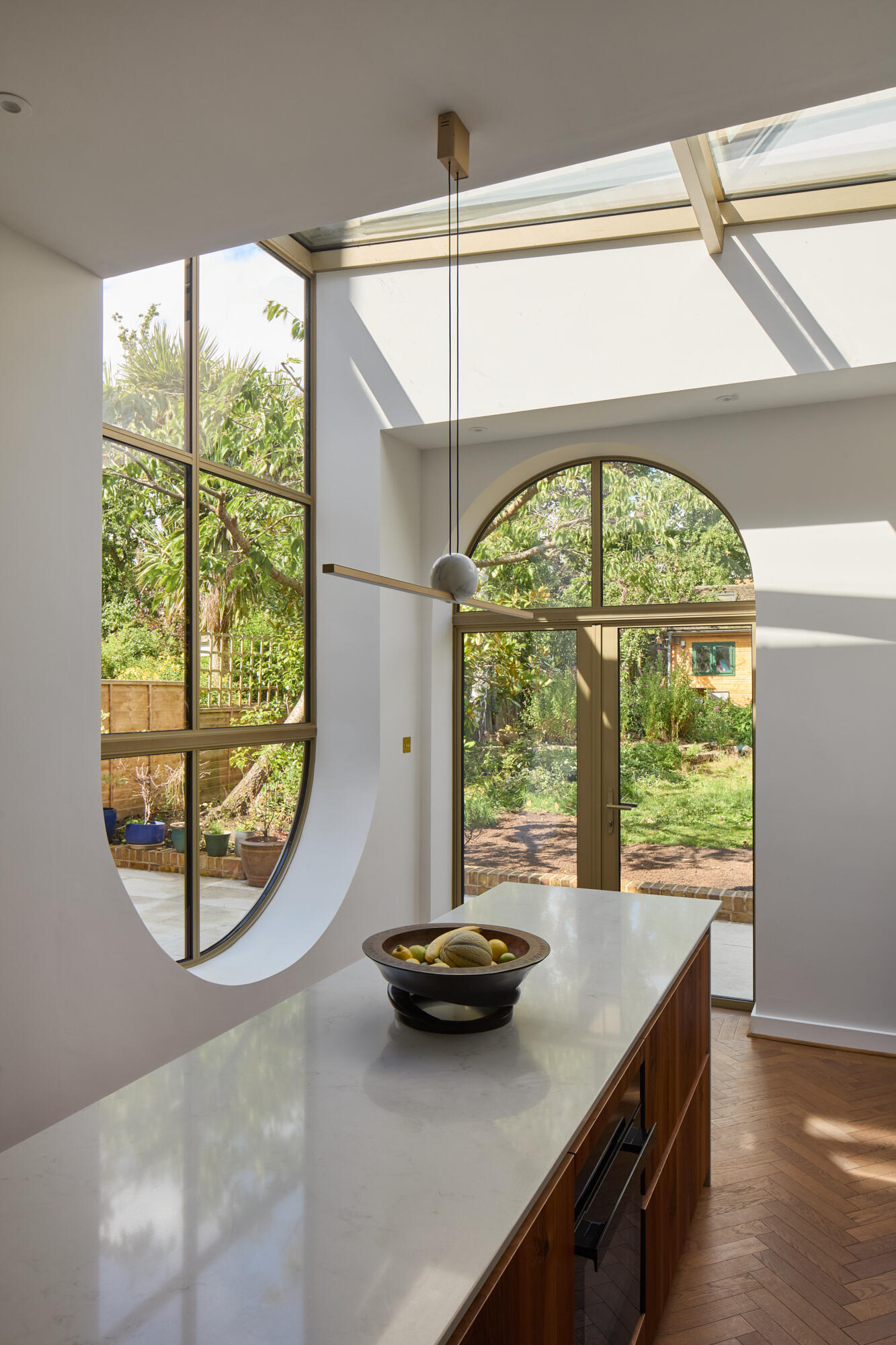 In the kitchen of Perry Vale House, inverted arches and a wraparound glass roof fill the space with natural light. Soft grey cabinetry, marble worktops, and timber tones create a warm, modern feel that stays true to the home’s character.