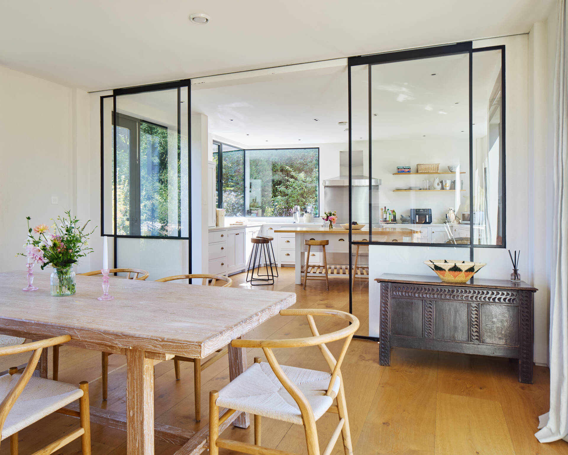 Behind the living room, the dining area introduces a soft, natural warmth with its pale timber table and calming palette. Large sliding glass panels open the space to the kitchen.
The kitchen shifts the mood to something brighter and more active. White cabinetry, open shelving, and generous windows bring in daylight and views of the garden. A central island with stools invites casual moments throughout the day.