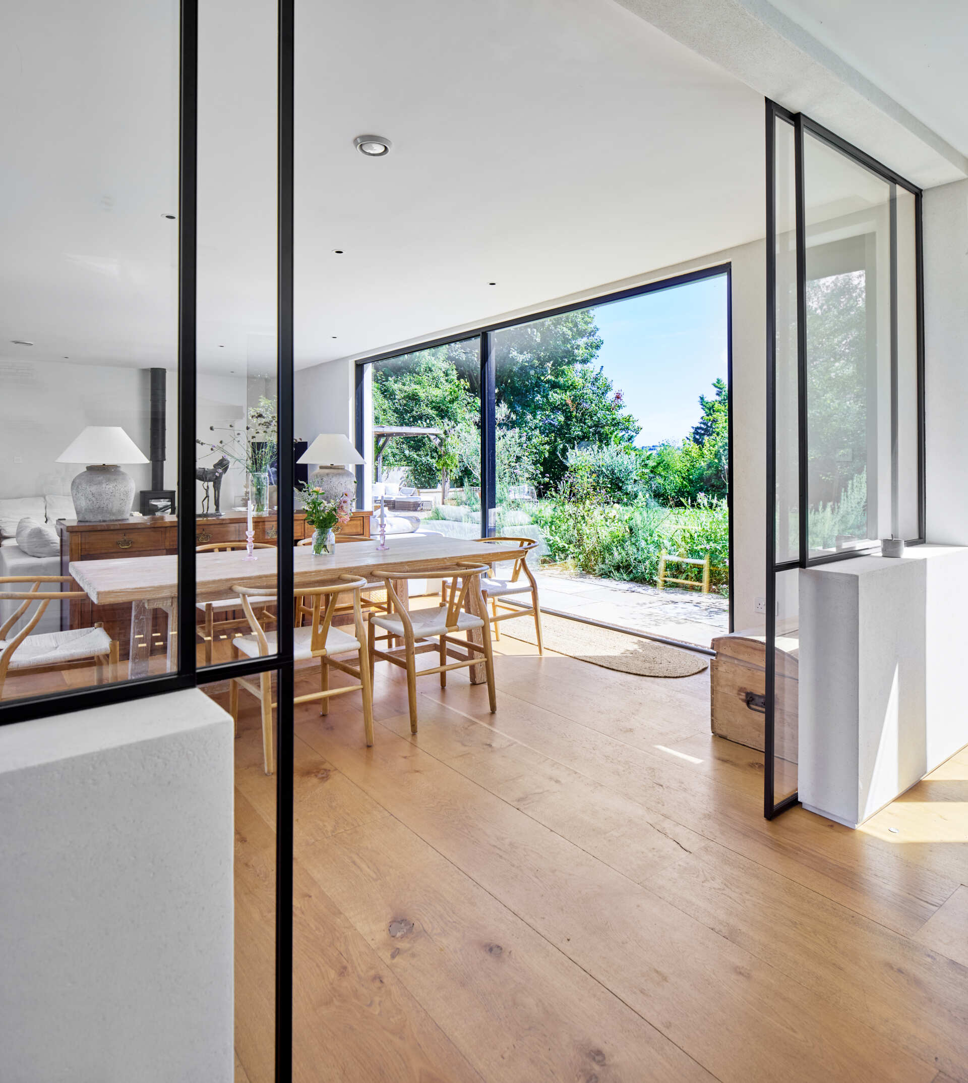 Behind the living room, the dining area introduces a soft, natural warmth with its pale timber table and calming palette. Large sliding glass panels open the space to the kitchen.
The kitchen shifts the mood to something brighter and more active. White cabinetry, open shelving, and generous windows bring in daylight and views of the garden. A central island with stools invites casual moments throughout the day.