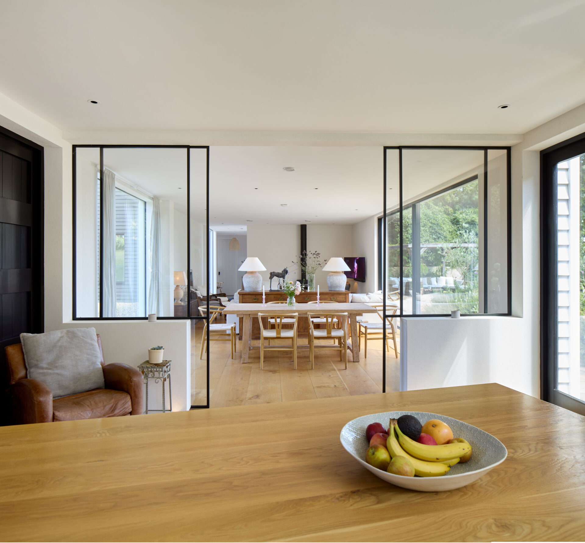 Behind the living room, the dining area introduces a soft, natural warmth with its pale timber table and calming palette. Large sliding glass panels open the space to the kitchen.
The kitchen shifts the mood to something brighter and more active. White cabinetry, open shelving, and generous windows bring in daylight and views of the garden. A central island with stools invites casual moments throughout the day.