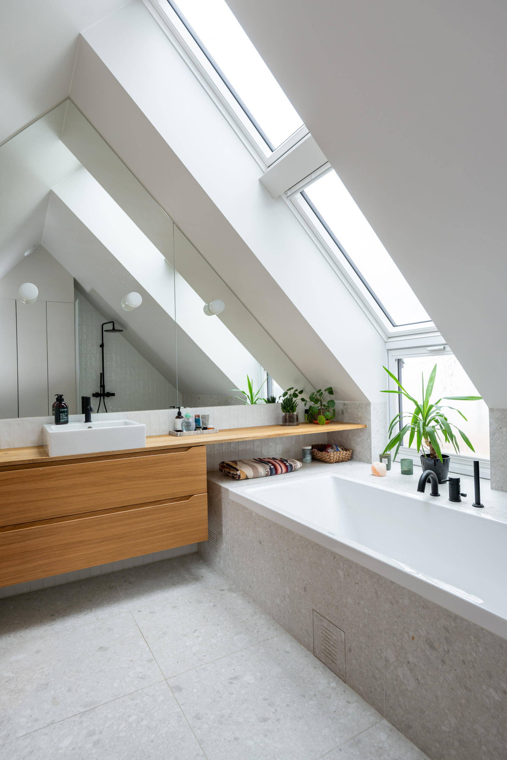 In the bathroom, natural light floods the room, while the mirrors follow the angled roof line, and a wood vanity floats above the tiled floor.