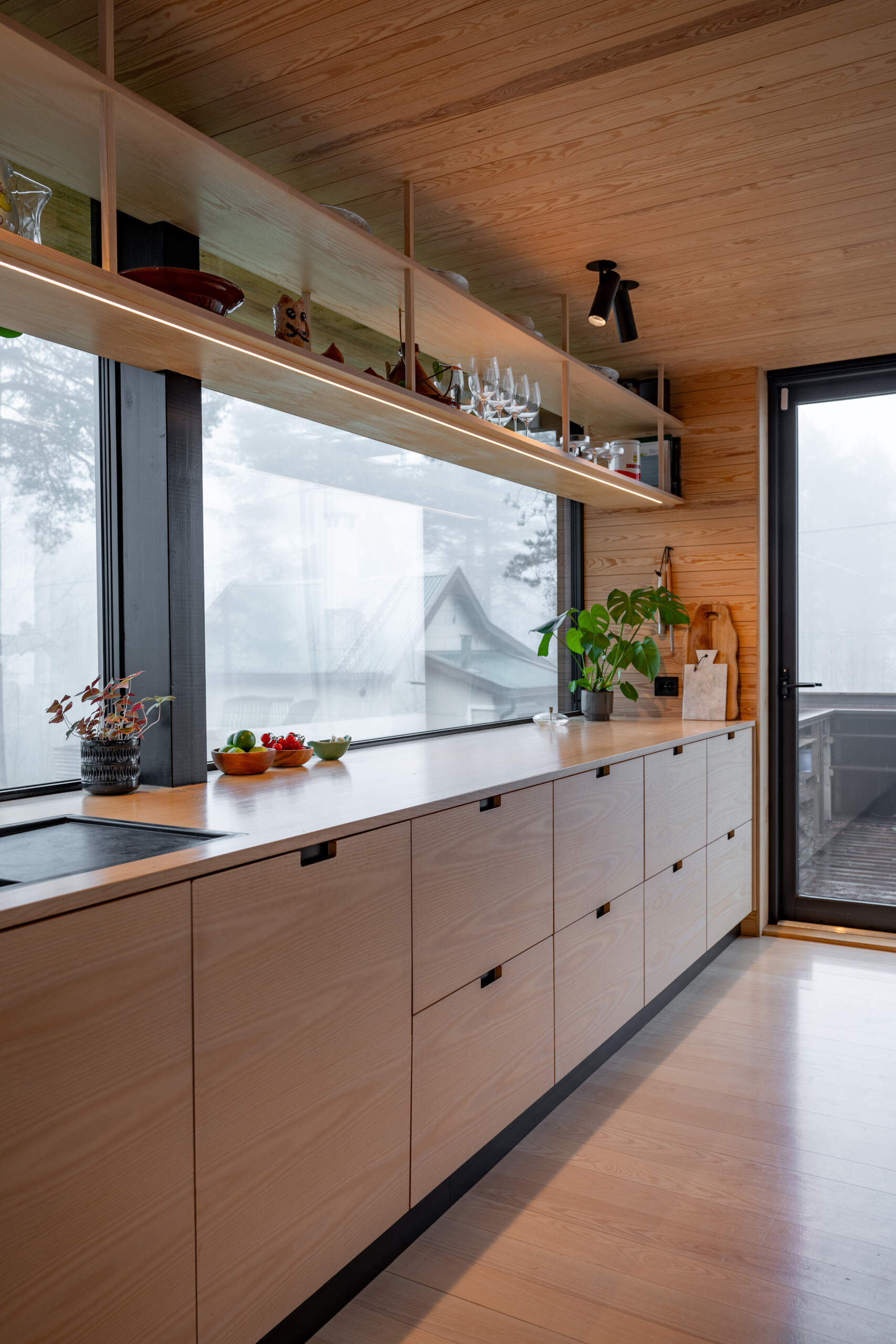 The kitchen, with hanging open shelving, is designed for efficiency and adaptability. Built-in storage and practical work surfaces ensure that daily routines are simple, while the timber finishes maintain warmth and texture.