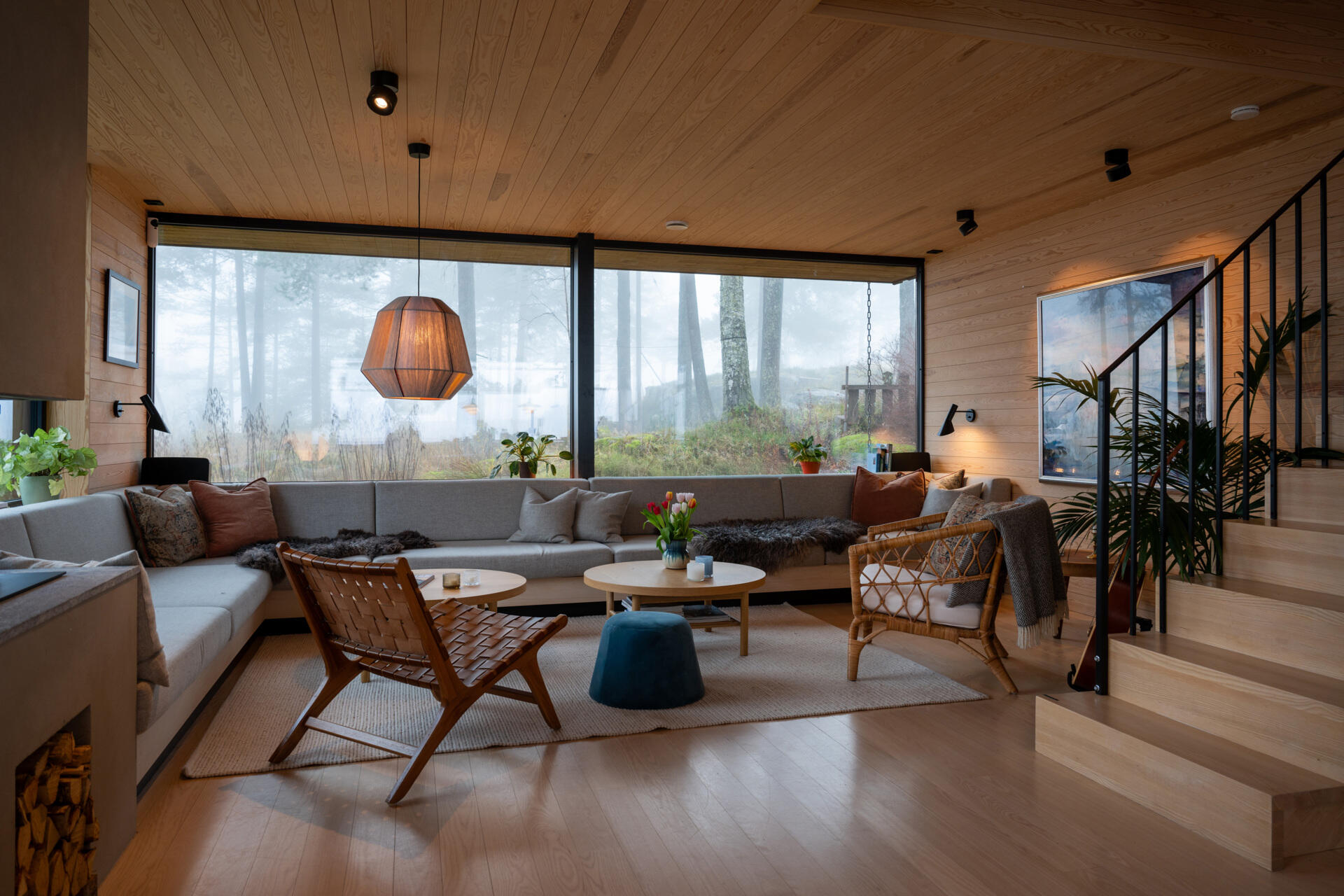 The living room, with its corner couch and fireplace, flows onto a timber deck, blending indoor comfort with the forest beyond. Sliding doors and large windows fill the space with daylight and frame views of the pine trees.