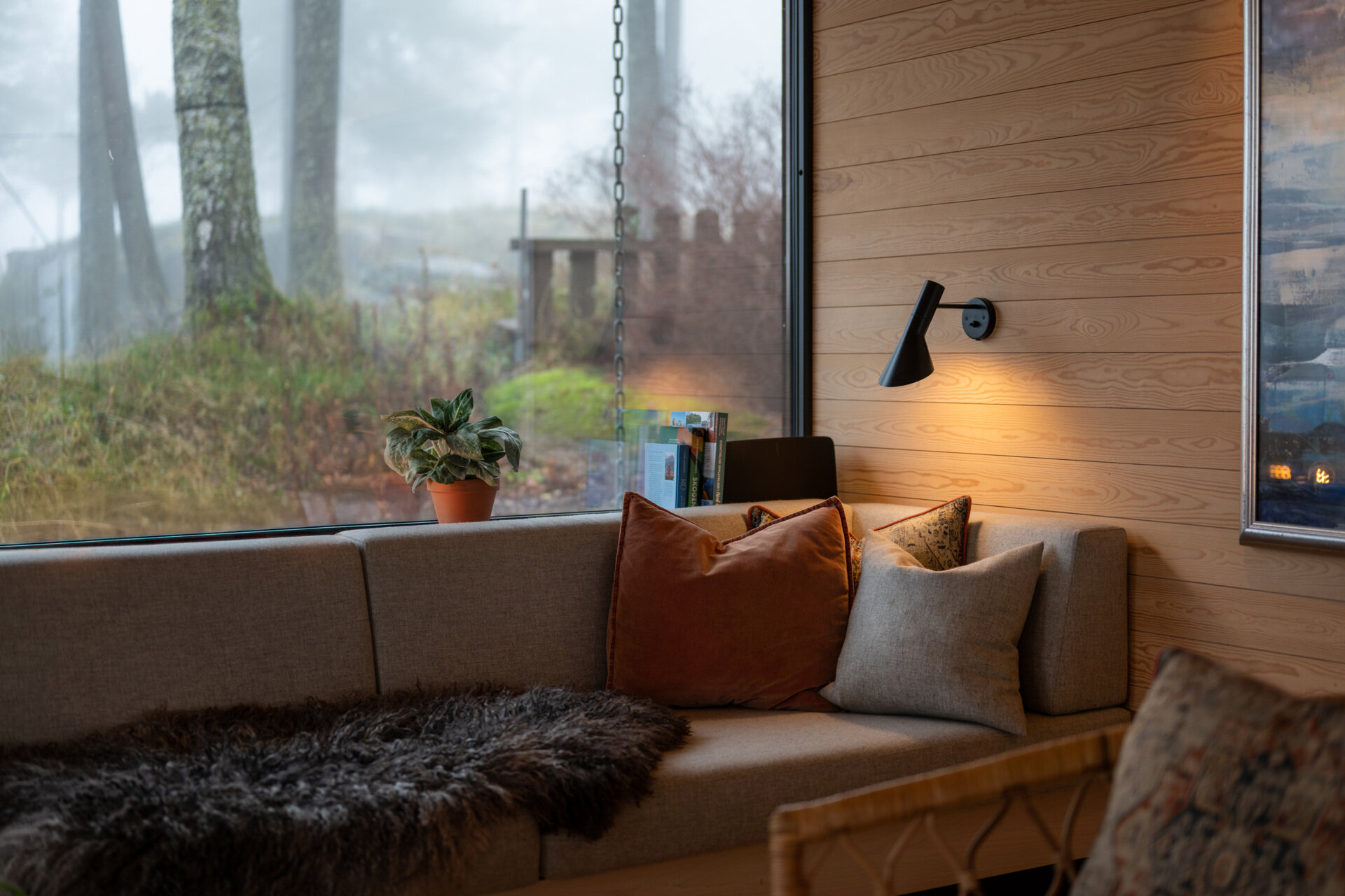 The living room, with its corner couch and fireplace, flows onto a timber deck, blending indoor comfort with the forest beyond. Sliding doors and large windows fill the space with daylight and frame views of the pine trees.