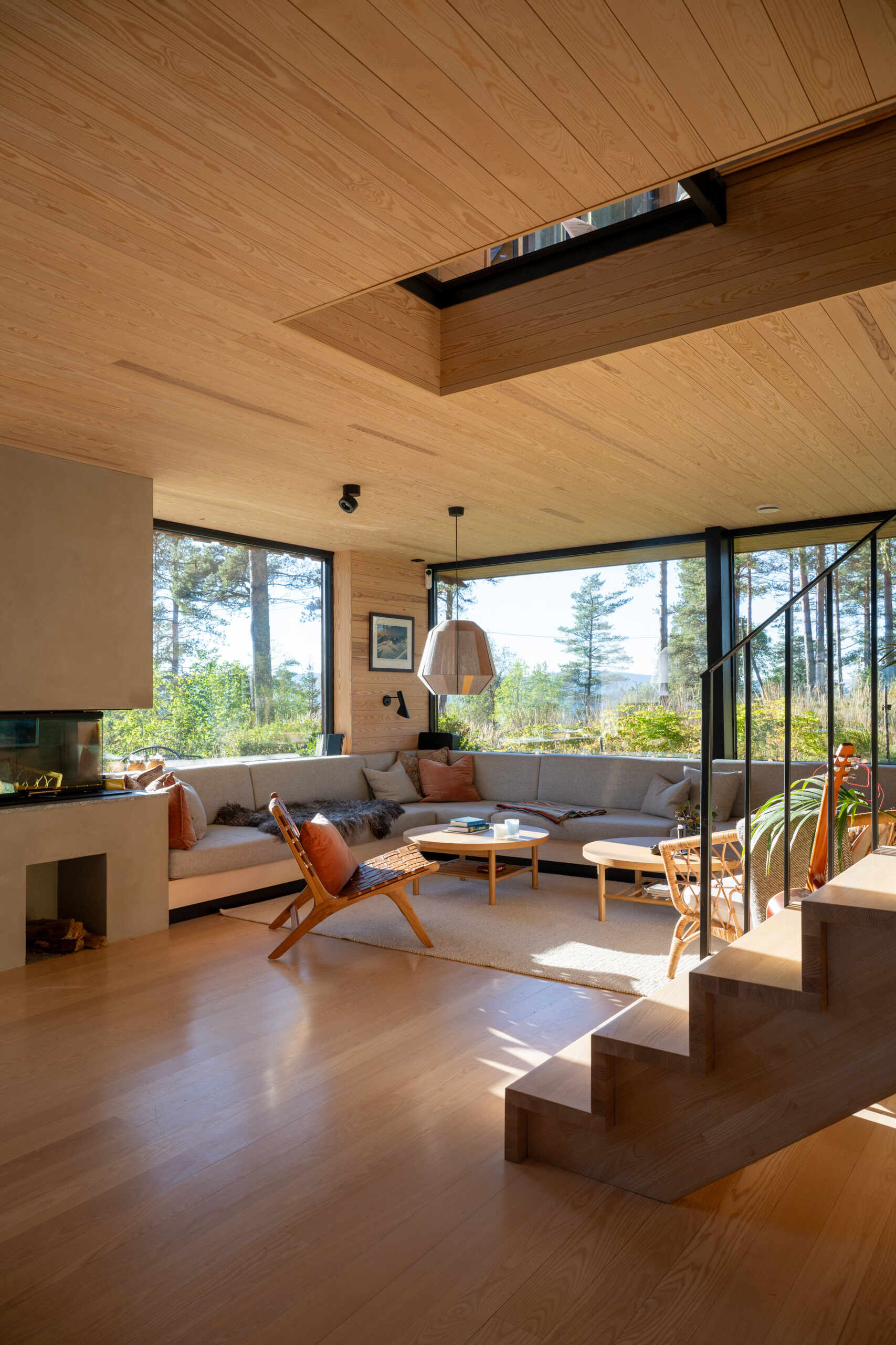 The living room, with its corner couch and fireplace, flows onto a timber deck, blending indoor comfort with the forest beyond. Sliding doors and large windows fill the space with daylight and frame views of the pine trees.