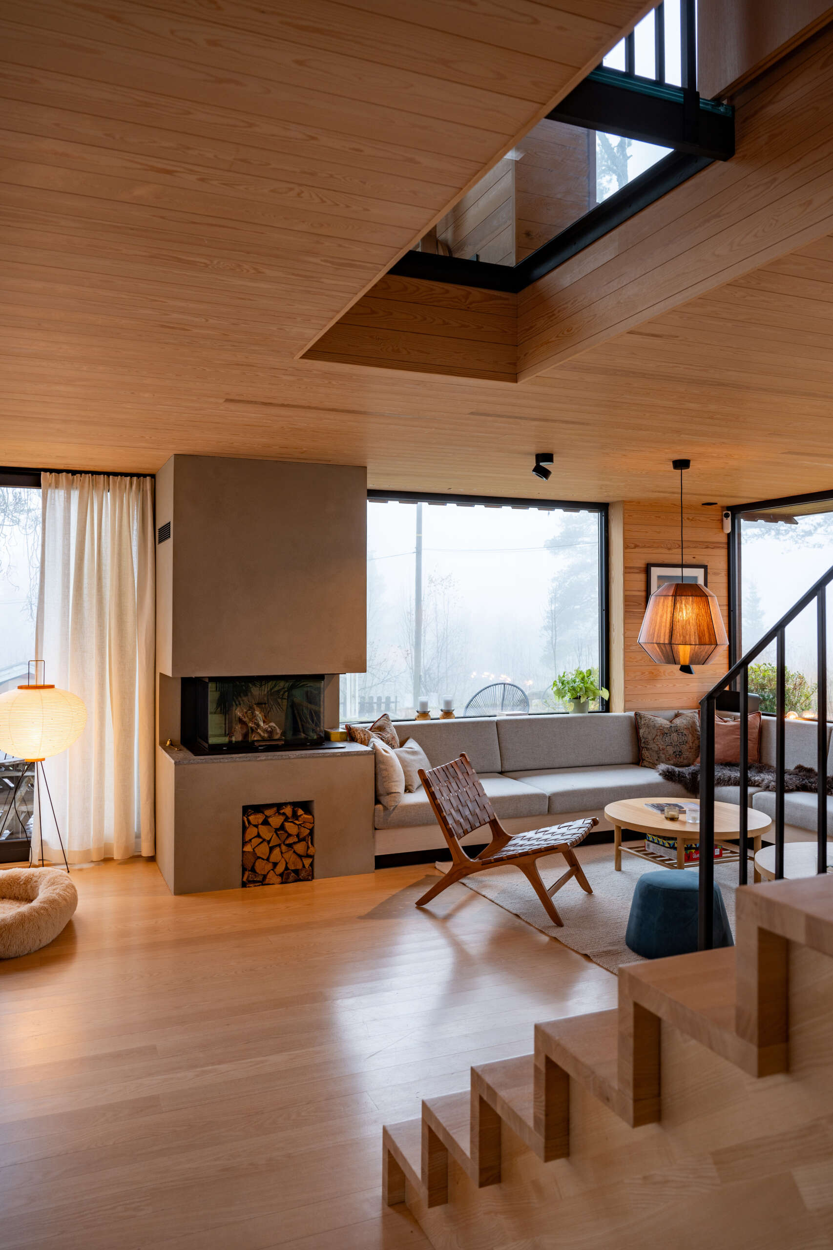 The living room, with its corner couch and fireplace, flows onto a timber deck, blending indoor comfort with the forest beyond. Sliding doors and large windows fill the space with daylight and frame views of the pine trees.