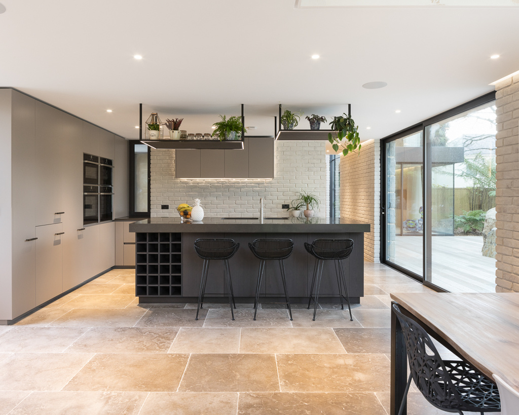 At the heart of the home, the kitchen feels modern yet understated. A dark grey island contrasts gently with the light grey cabinetry, while stone flooring grounds the space.