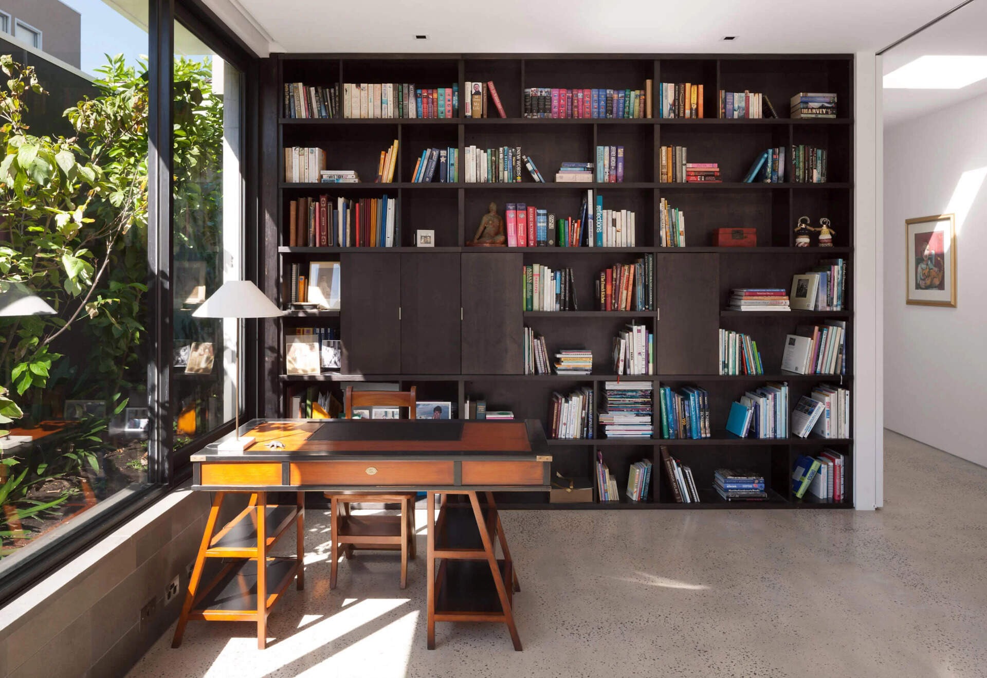 Somewhat concealed behind the courtyard plants is a door that leads to the office. Inside, floor to ceiling wood shelving lines the walls, while a desk sits by the window. It is a compact but peaceful corner designed for work, reading or study with a view of the courtyard.