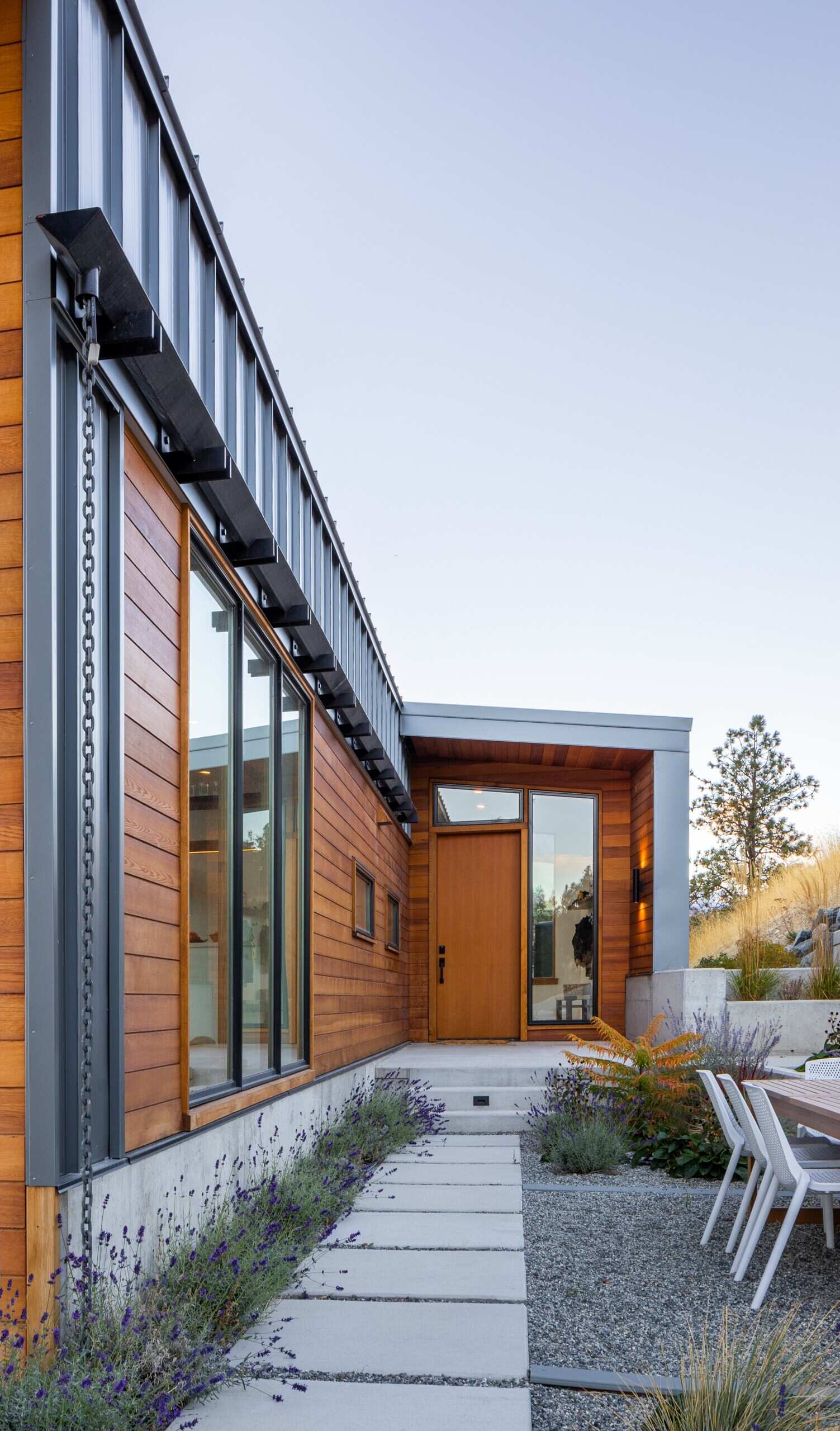 Una entrada tranquila y acogedora enmarcada por ventanas altas, revestimiento de cedro y plantas suaves lo conduce hacia la puerta principal de madera.