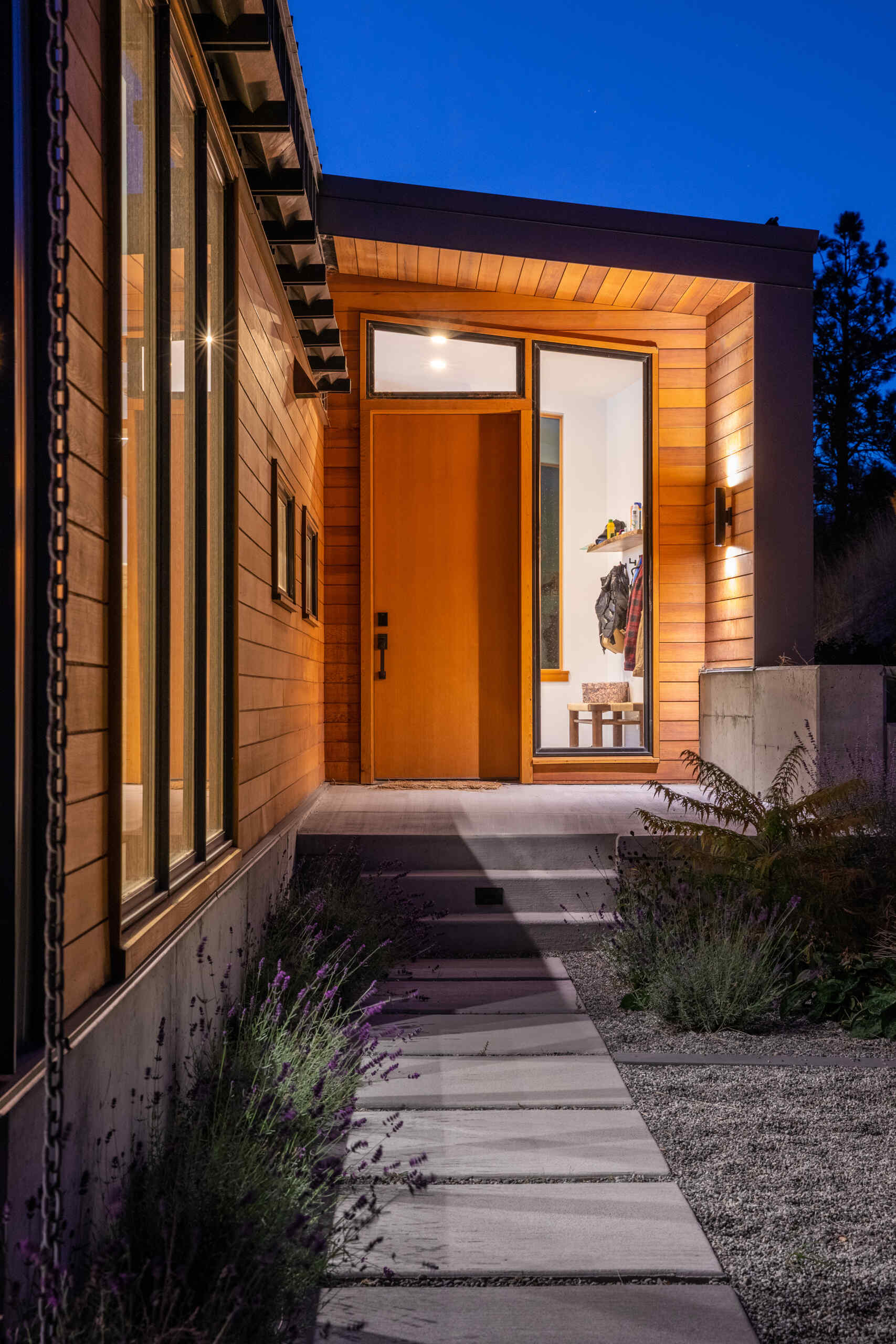 Una entrada tranquila y acogedora enmarcada por ventanas altas, revestimiento de cedro y plantas suaves lo conduce hacia la puerta principal de madera.