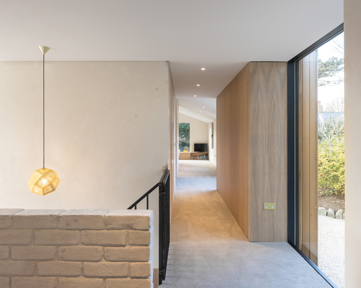 A bespoke raw steel staircase connects the two levels, sitting beside a wall of locally reclaimed brick coated in lime slurry. The contrast between metal and masonry adds texture to the journey upward, while soft light filters through from the double-height glazing that links both wings of the house.