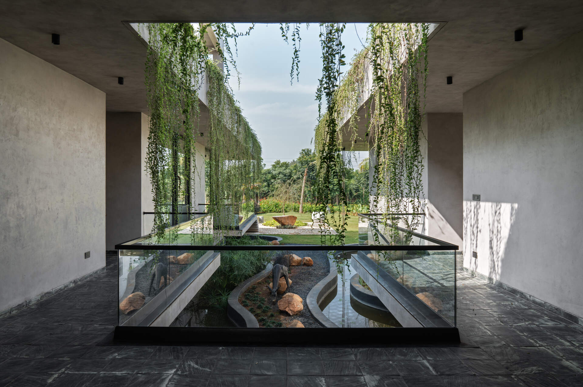 An outdoor walkway opens to a view of the level below. From this elevated path, the floating plinth, water body, and landscaped pockets become part of a larger composition.
