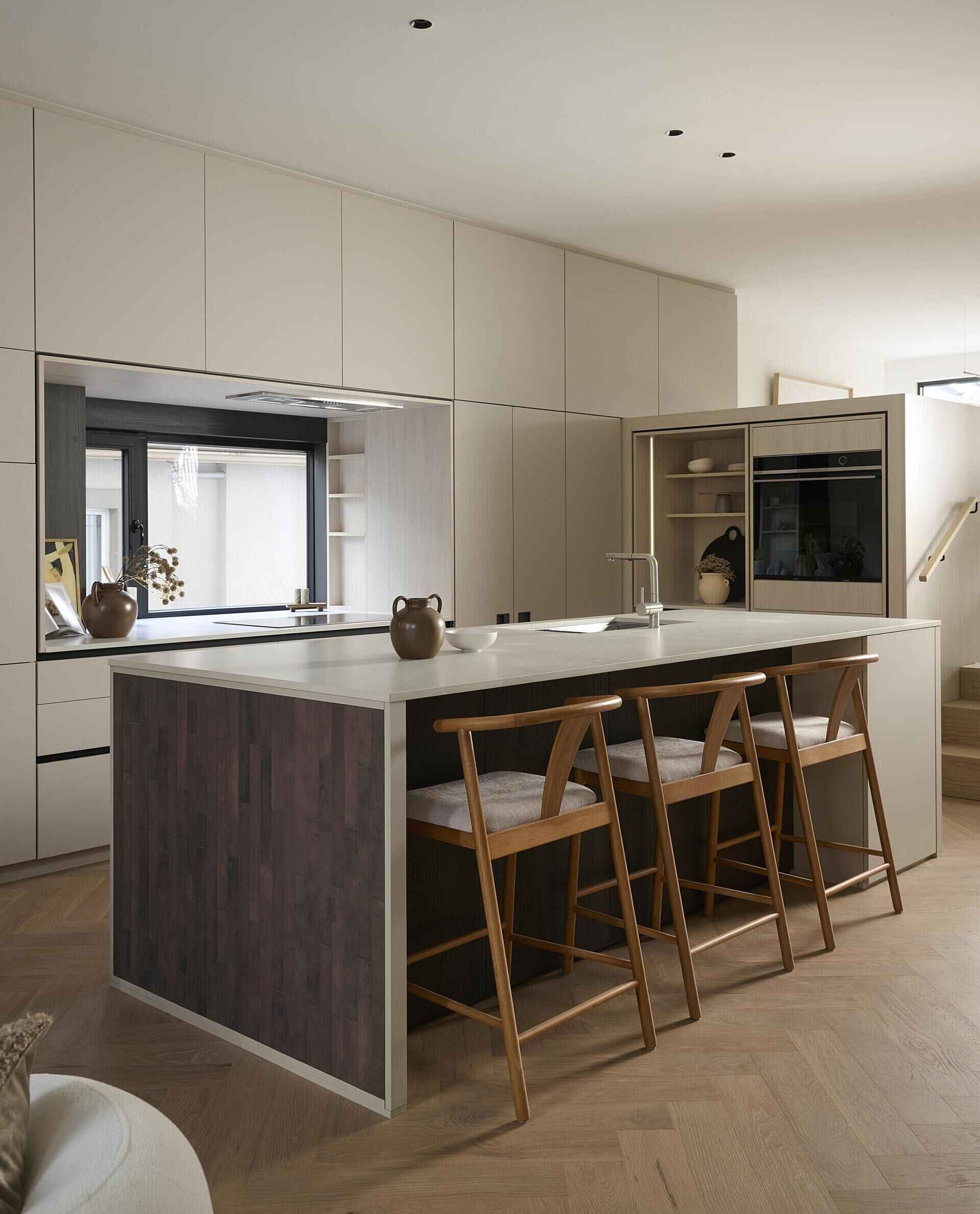 At the centre of this modern home, the kitchen sits against the stairwell. A large island in concrete toned Caesarstone pairs with light grey and whitewashed oak cabinetry trimmed in black. The island’s red toned brick end gables add gentle warmth. A full width window replaces a traditional backsplash, pulling natural light and surprising depth into the room.