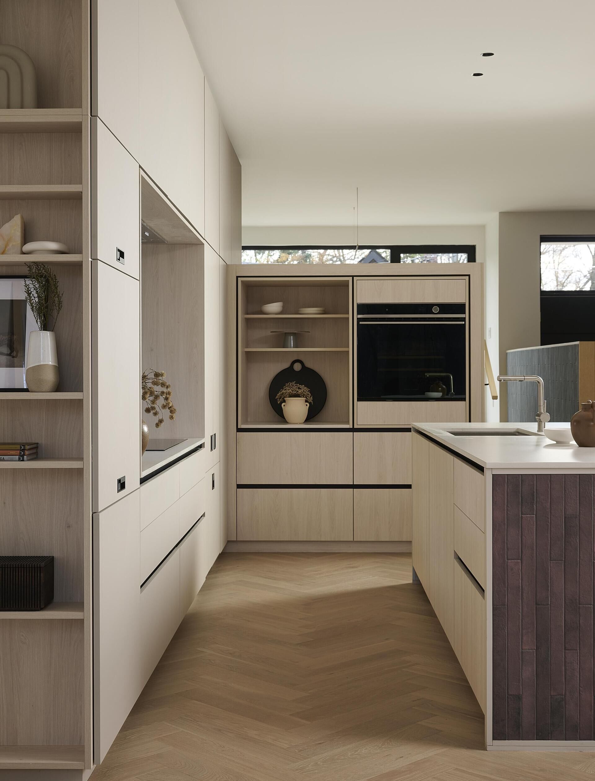 At the centre of this modern home, the kitchen sits against the stairwell. A large island in concrete toned Caesarstone pairs with light grey and whitewashed oak cabinetry trimmed in black. The island’s red toned brick end gables add gentle warmth. A full width window replaces a traditional backsplash, pulling natural light and surprising depth into the room.