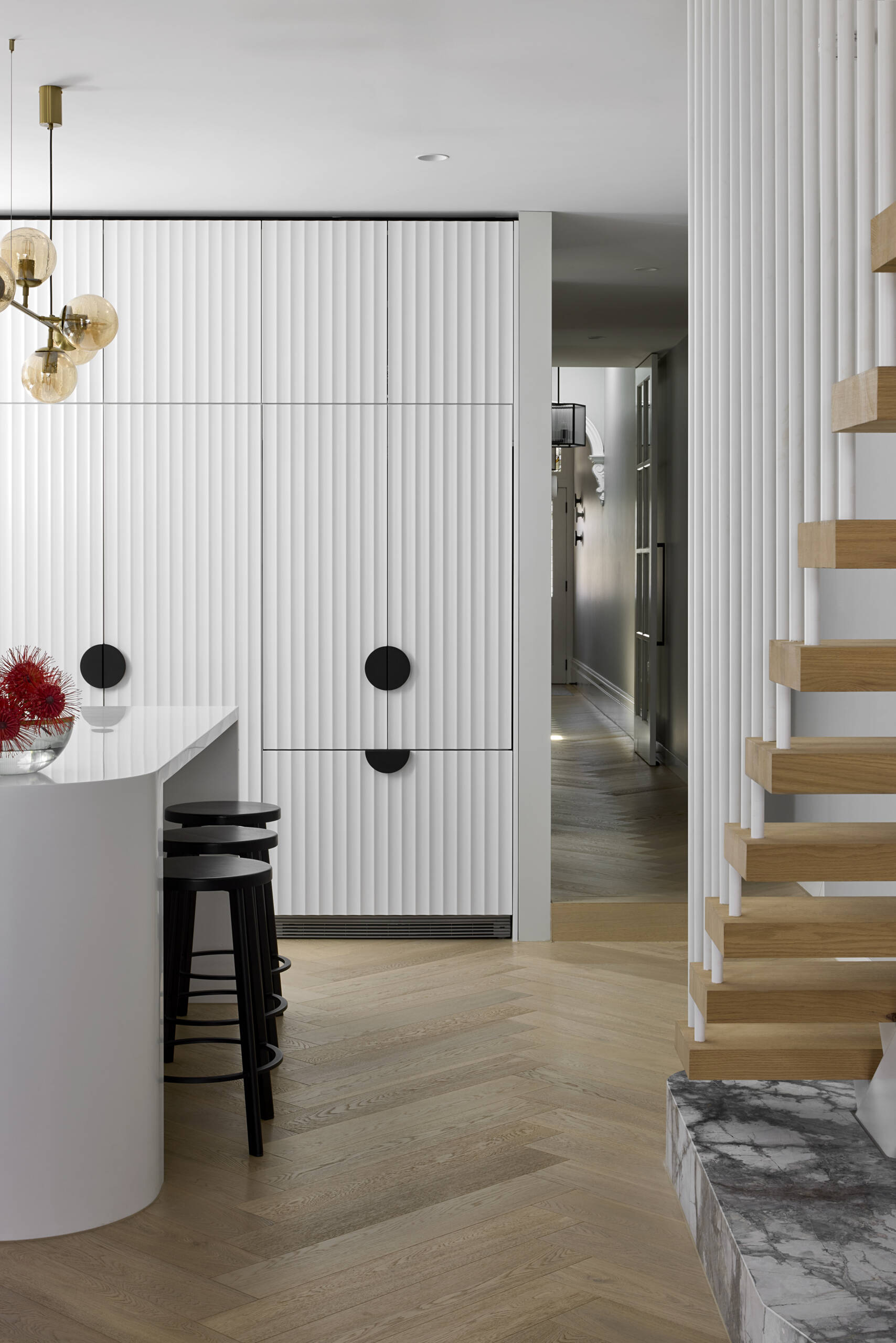 In this modern kitchen, a dramatic waterfall skylight pours daylight into what was once a dark corner. A curved white island and fluted cabinetry create a clean, sculptural base for the bold grey and white marble that lines the walls and splashback. Warm herringbone floors anchor the space, while black stools, fittings, and soft golden globes add balance. The island becomes the natural point for daily gathering.
