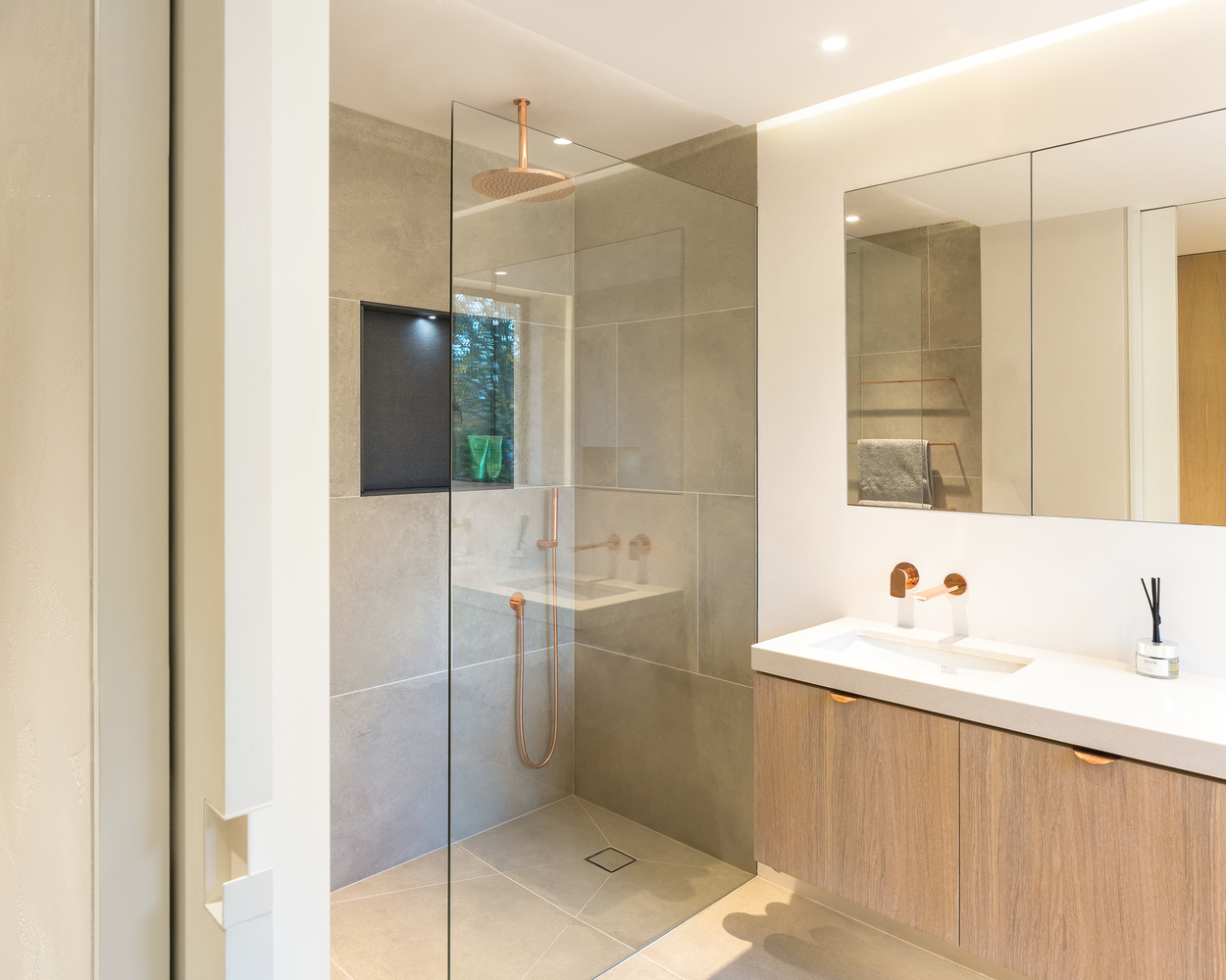 In the ensuite bathroom, a walk-in shower with a rainfall head and concealed lighting creates a spa-like calm. Subtle uplighting above the mirror enhances the gentle tones of the materials, while a skylight above the toilet area allows natural light to pour in, making even the smallest corner feel open.