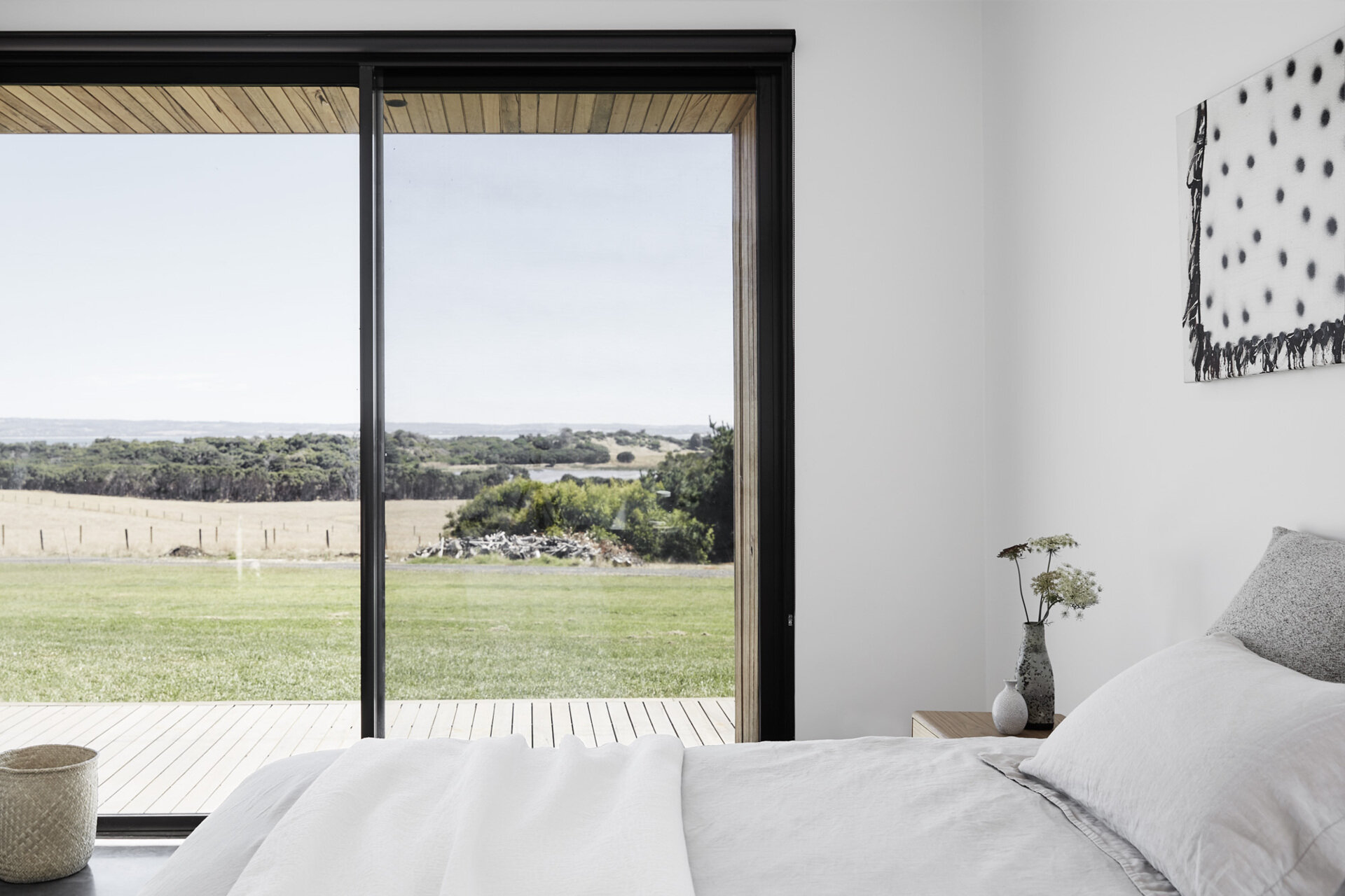 A calm bedroom with simple furnishings opens to the deck through a sliding glass door that captures fresh air and coastal views.