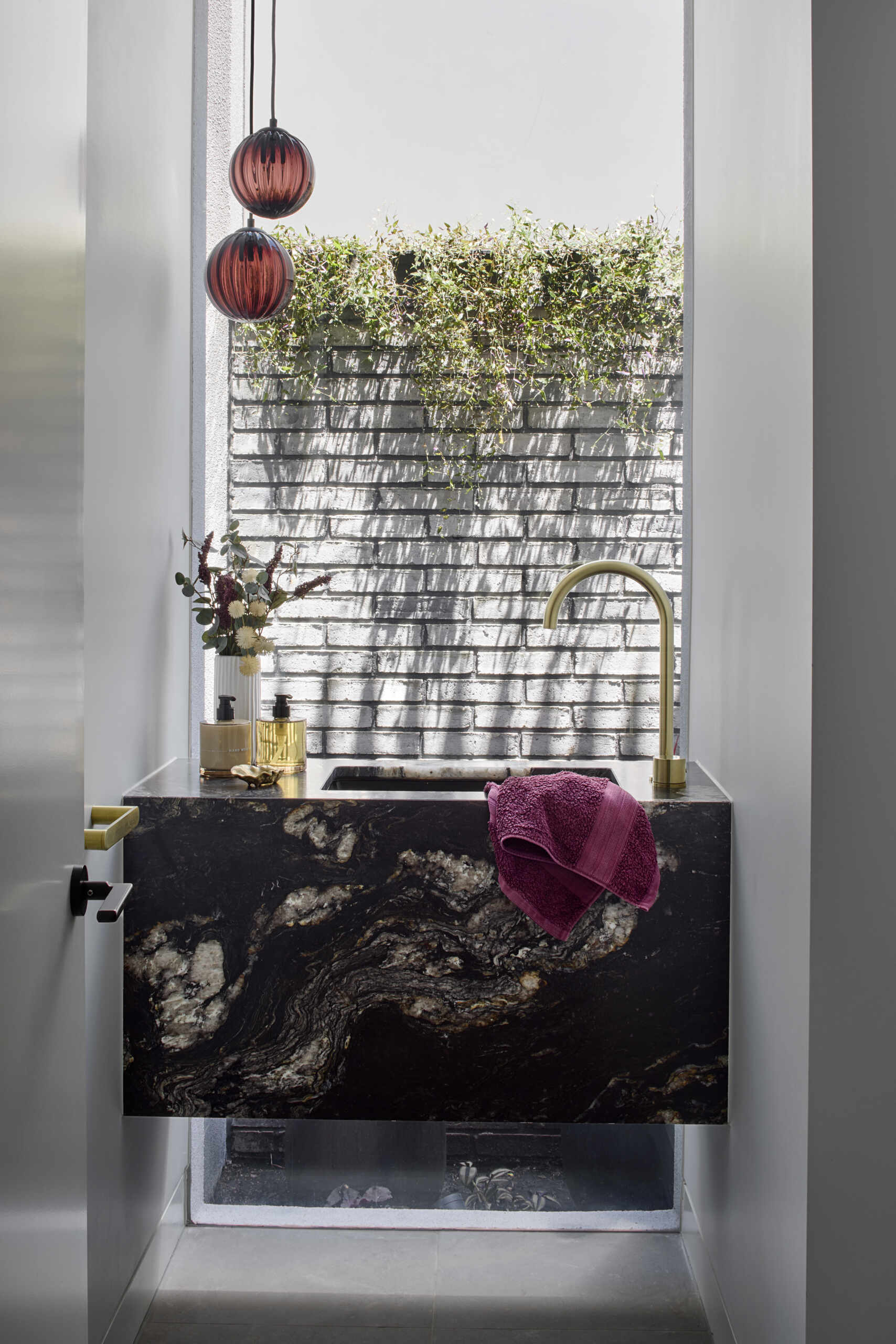 The powder room feels like a small highlight within the home. Views through a window showcase the sunlight as it falls across a brick wall softened with greenery. A floating black stone vanity marked with gold and cream veining stands out, supported by warm brass fittings. Berry toned towels and ribbed glass pendants bring depth to the compact room.