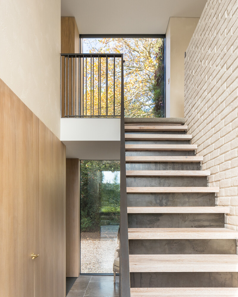 A bespoke raw steel staircase connects the two levels, sitting beside a wall of locally reclaimed brick coated in lime slurry. The contrast between metal and masonry adds texture to the journey upward, while soft light filters through from the double-height glazing that links both wings of the house.
