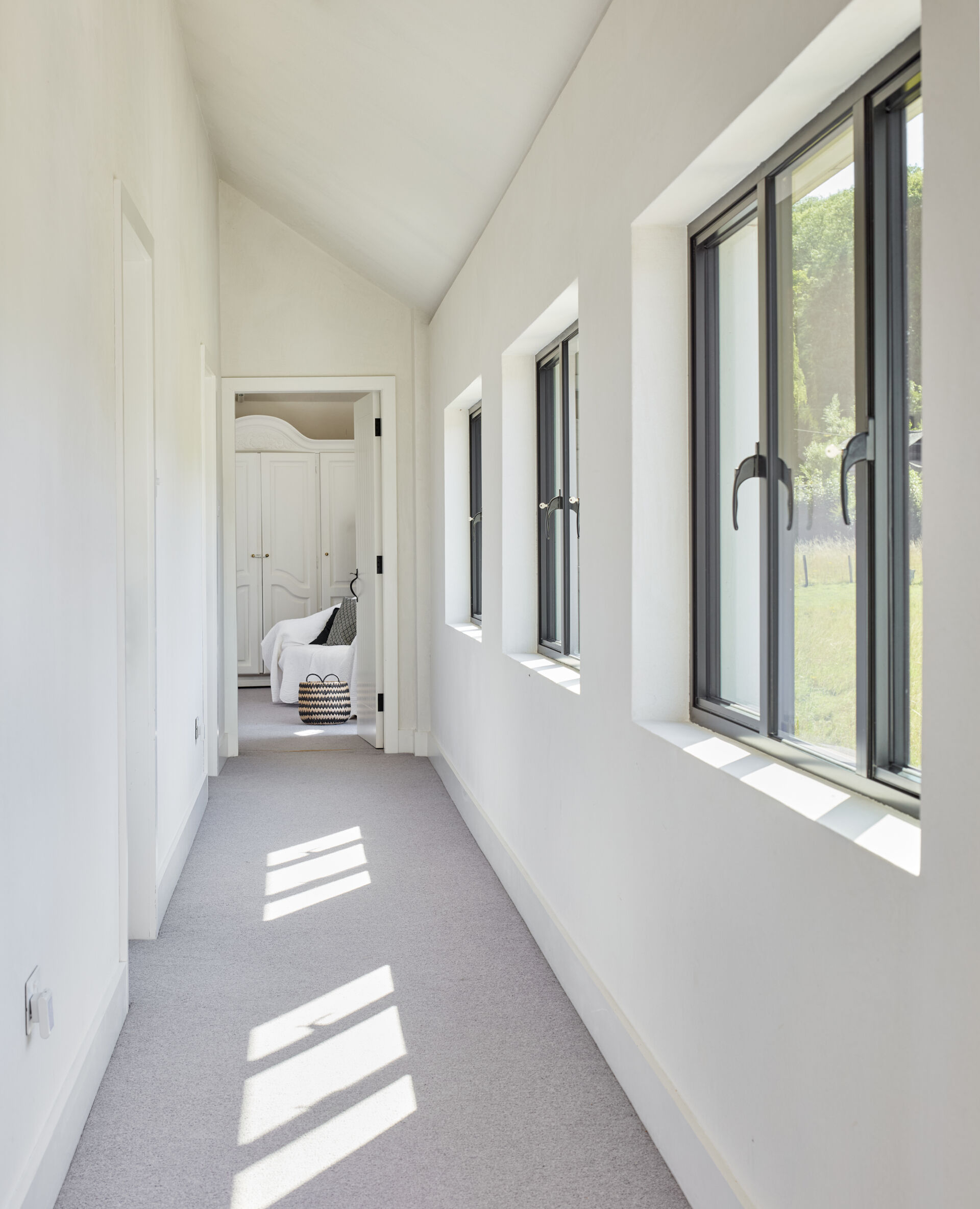 The hallway stretches quietly along a row of deep set windows that catch the light throughout the day. Soft carpet keeps the space calm and muted, guiding you toward the bedroom at the far end. The simplicity of the white walls and angled ceiling gives the passage a gentle, airy feel.