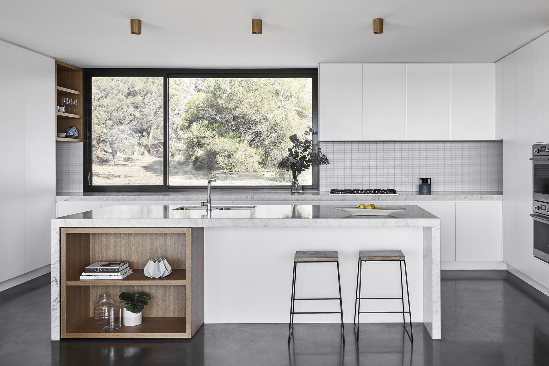 Minimalist white cabinetry in the kitchen is paired with exposed wood shelving, creating a clean look that stands out against the dark window frames and flooring.