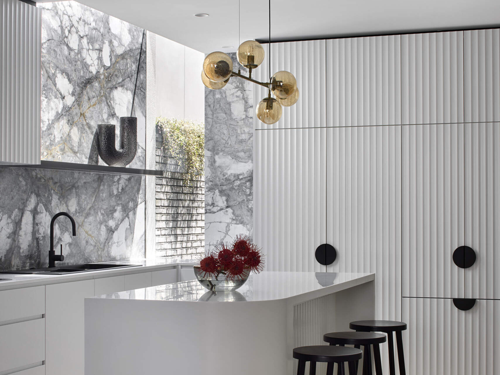 In this modern kitchen, a dramatic waterfall skylight pours daylight into what was once a dark corner. A curved white island and fluted cabinetry create a clean, sculptural base for the bold grey and white marble that lines the walls and splashback. Warm herringbone floors anchor the space, while black stools, fittings, and soft golden globes add balance. The island becomes the natural point for daily gathering.
