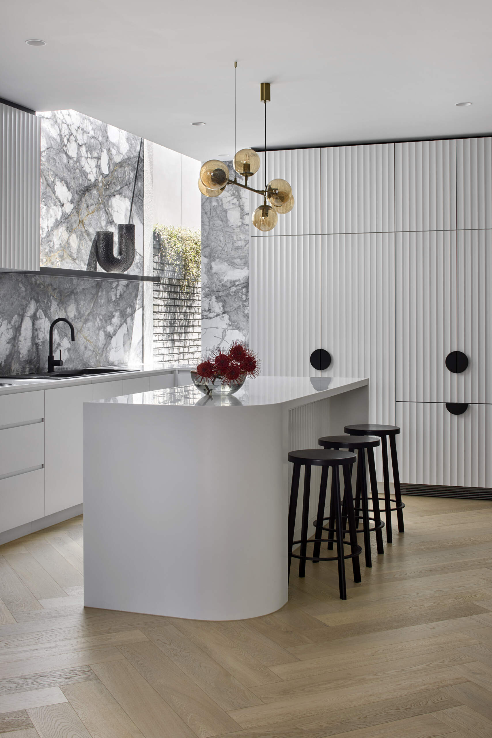 In this modern kitchen, a dramatic waterfall skylight pours daylight into what was once a dark corner. A curved white island and fluted cabinetry create a clean, sculptural base for the bold grey and white marble that lines the walls and splashback. Warm herringbone floors anchor the space, while black stools, fittings, and soft golden globes add balance. The island becomes the natural point for daily gathering.