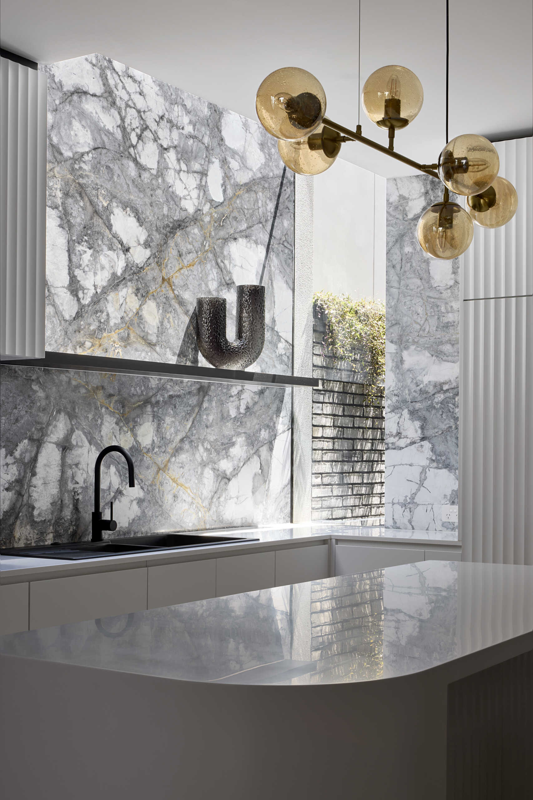 In this modern kitchen, a dramatic waterfall skylight pours daylight into what was once a dark corner. A curved white island and fluted cabinetry create a clean, sculptural base for the bold grey and white marble that lines the walls and splashback. Warm herringbone floors anchor the space, while black stools, fittings, and soft golden globes add balance. The island becomes the natural point for daily gathering.