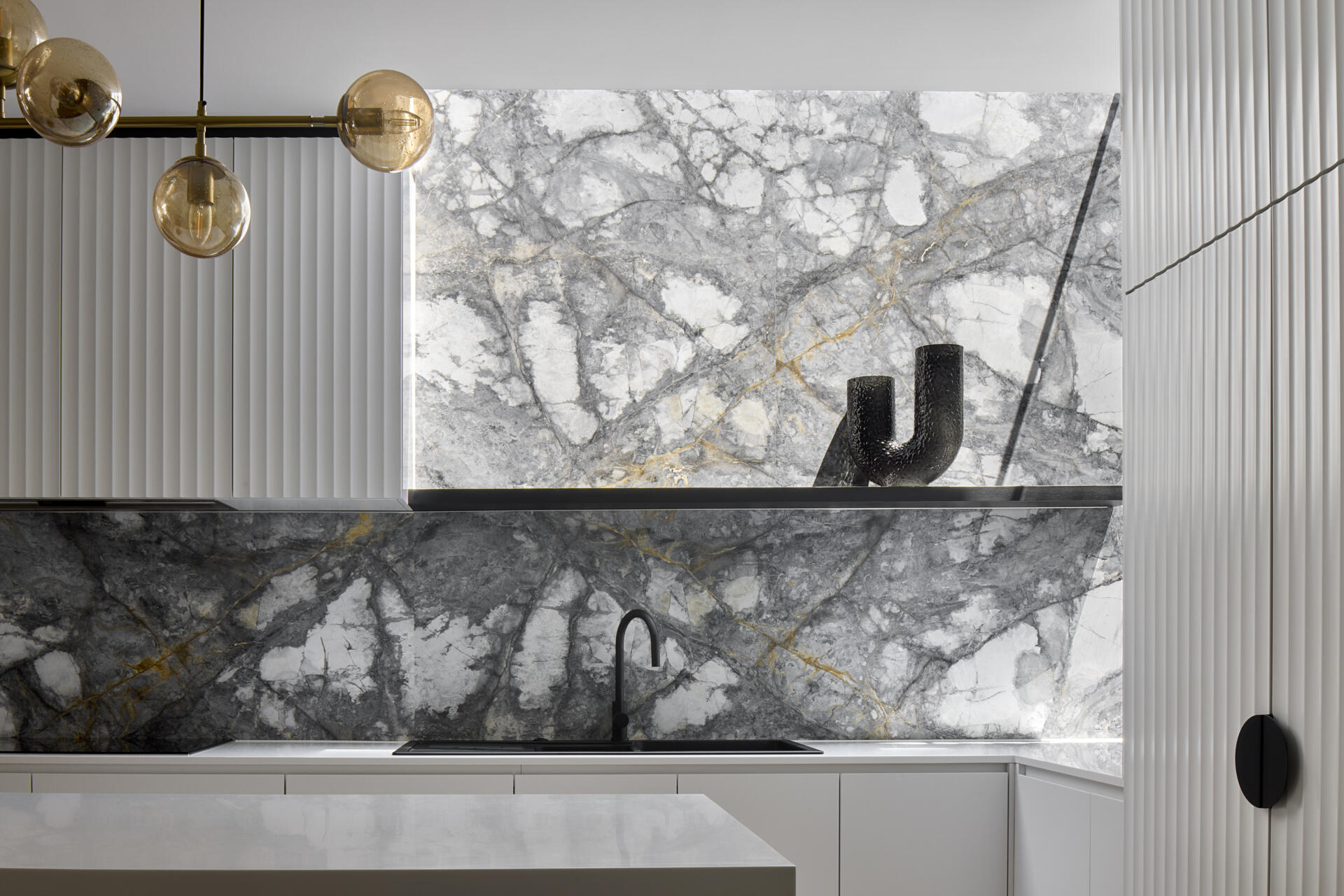 In this modern kitchen, a dramatic waterfall skylight pours daylight into what was once a dark corner. A curved white island and fluted cabinetry create a clean, sculptural base for the bold grey and white marble that lines the walls and splashback. Warm herringbone floors anchor the space, while black stools, fittings, and soft golden globes add balance. The island becomes the natural point for daily gathering.