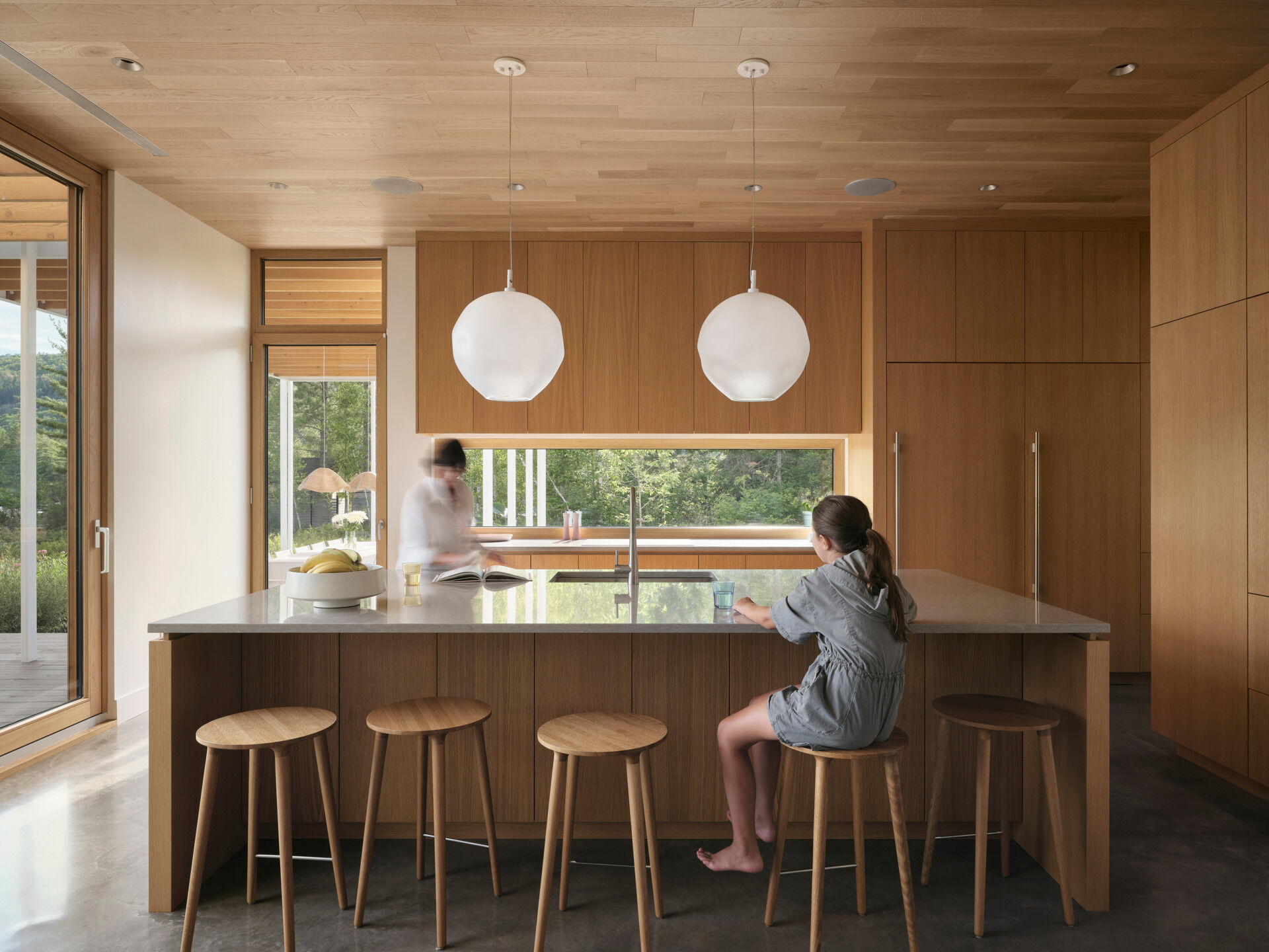 The dining and kitchen areas flow directly from the living room, continuing the soft neutral tones. Wood cabinetry, a wood dining table, and a large island keep it contemporary, while a window backsplash provides a view of the trees.