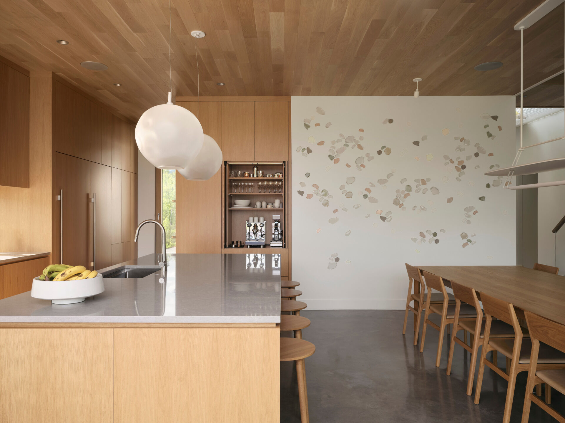 The dining and kitchen areas flow directly from the living room, continuing the soft neutral tones. Wood cabinetry, a wood dining table, and a large island keep it contemporary, while a window backsplash provides a view of the trees.