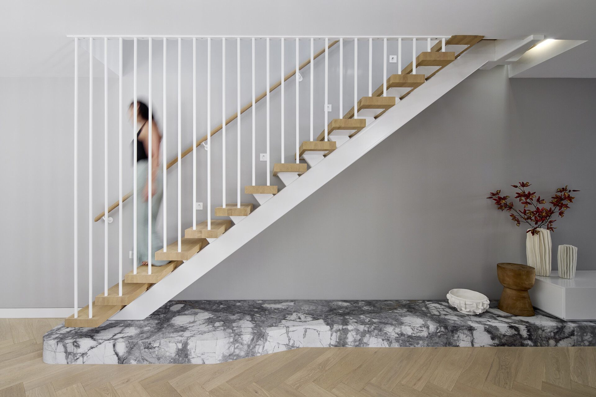 The staircase sits as a quiet feature, with pale wood treads floating above a marble platform. Slim white balusters keep the structure light. Grey walls and herringbone floors create a soft backdrop for simple pottery and branches that mark the transition between levels.