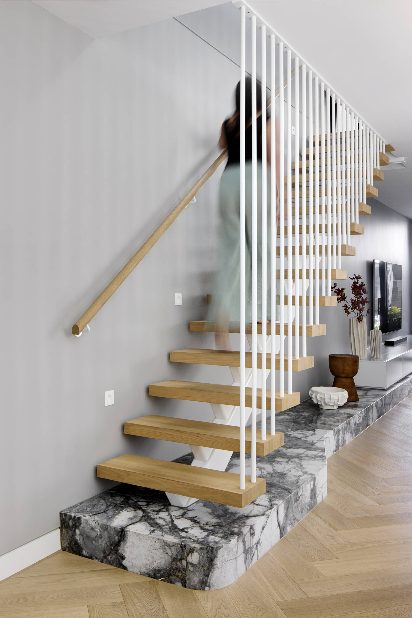 The staircase sits as a quiet feature, with pale wood treads floating above a marble platform. Slim white balusters keep the structure light. Grey walls and herringbone floors create a soft backdrop for simple pottery and branches that mark the transition between levels.